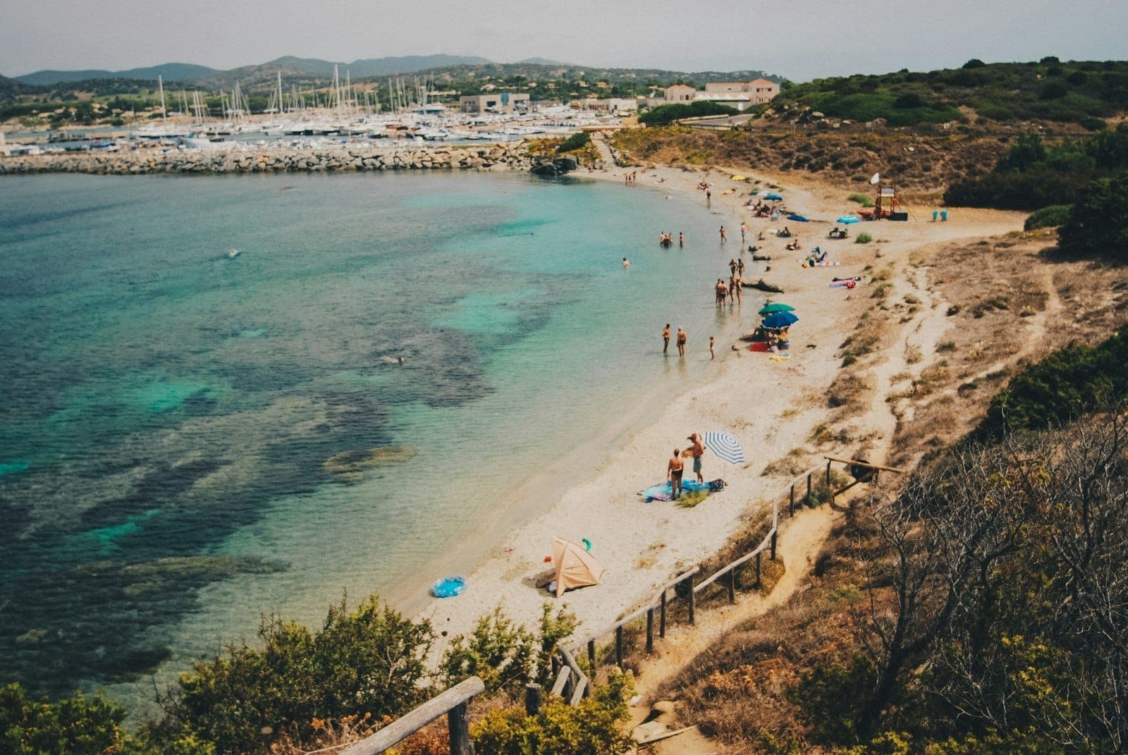 Una playa sarda con bañistas y sombrillas dispersas, agua turquesa cristalina, olas suaves y un puerto al fondo bajo cielos soleados.