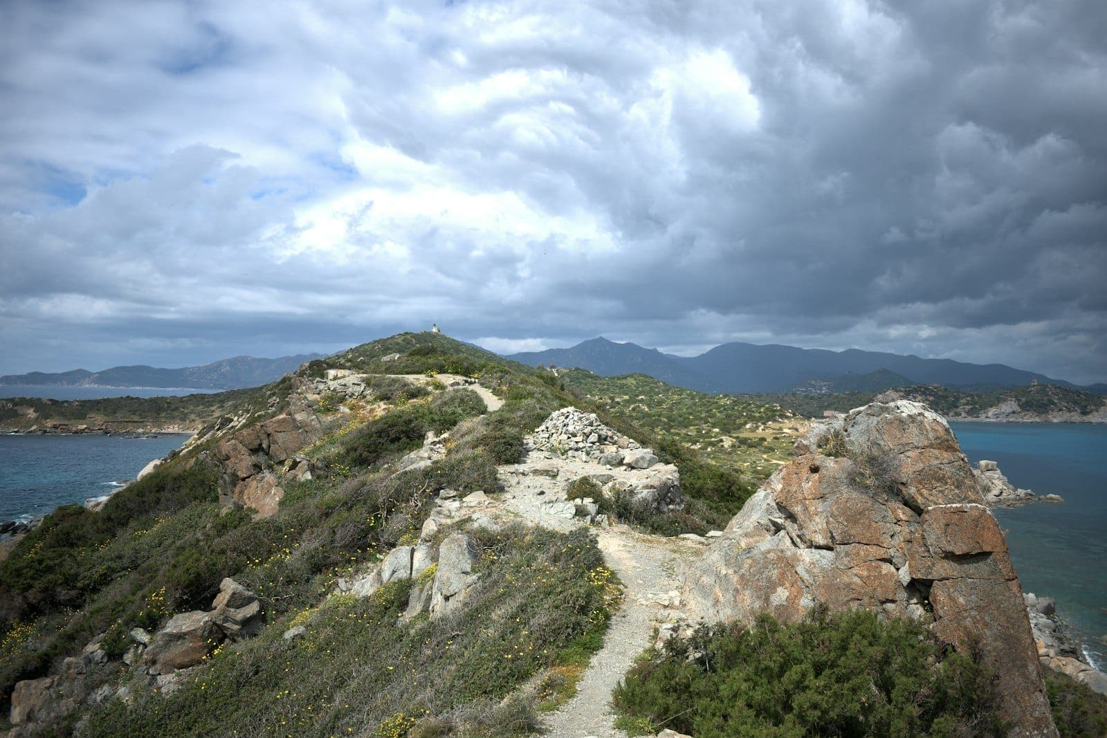 Sendero costero escarpado en lo alto de colinas rocosas en Cerdeña, con nubes dramáticas, arbustos verdes y un lejano mar azul.