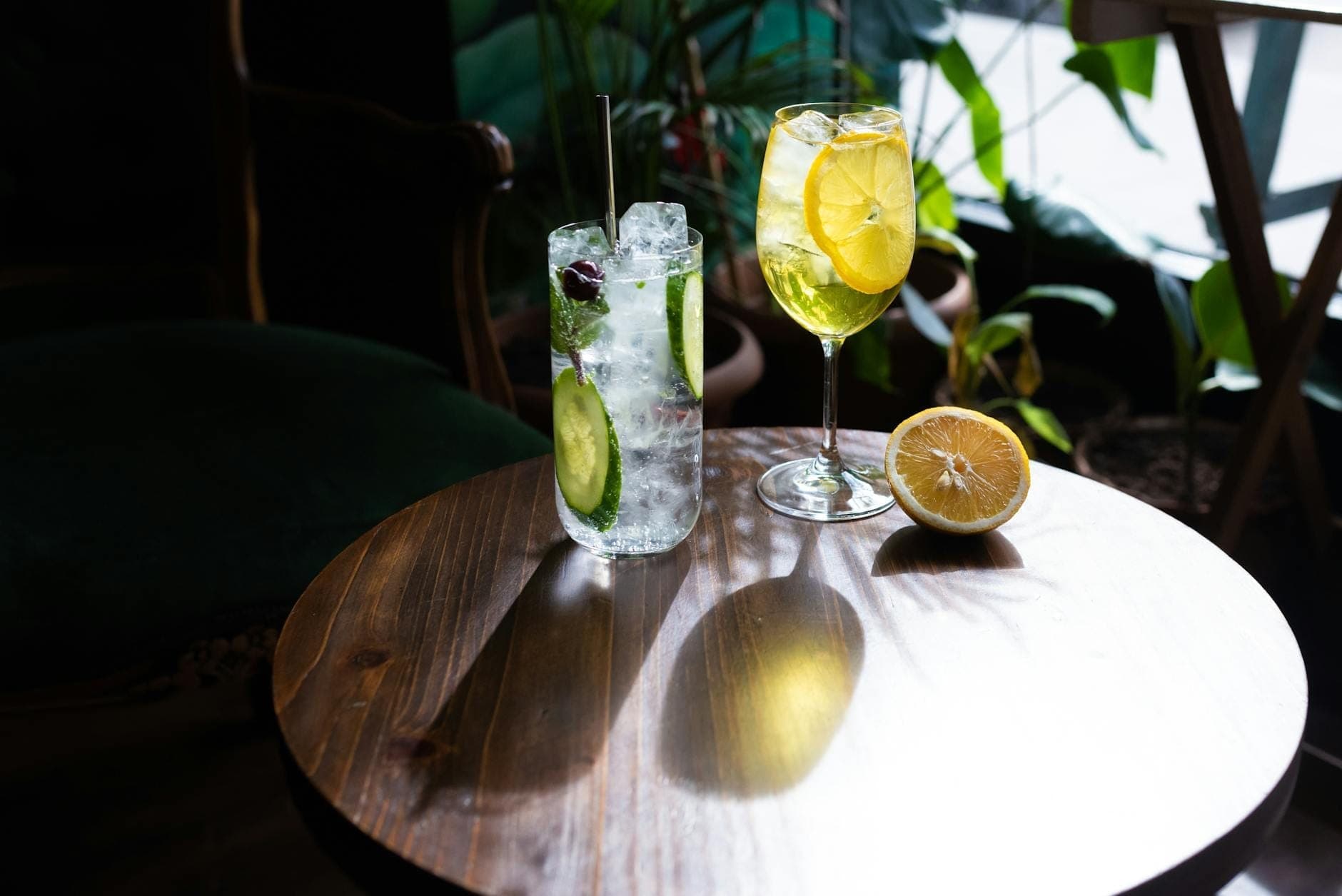 Dos elegantes bebidas sin alcohol sobre una mesa redonda de madera, decoradas con pepino fresco, cítricos y frutos rojos, con un fondo de cafetería luminoso y acogedor.