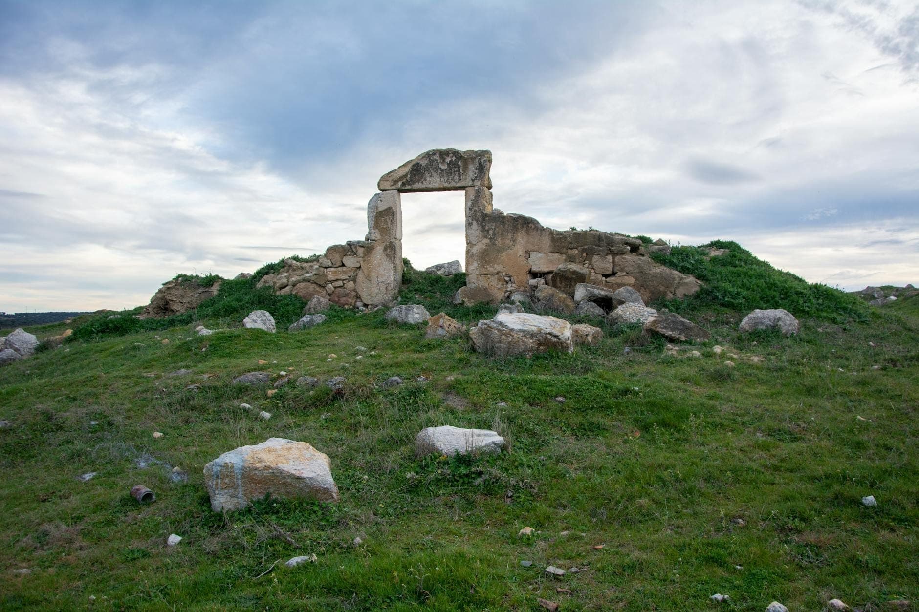 Antigua estructura funeraria de piedra con losas verticales y horizontales en un paisaje de hierba bajo un cielo nublado en Cerdeña.