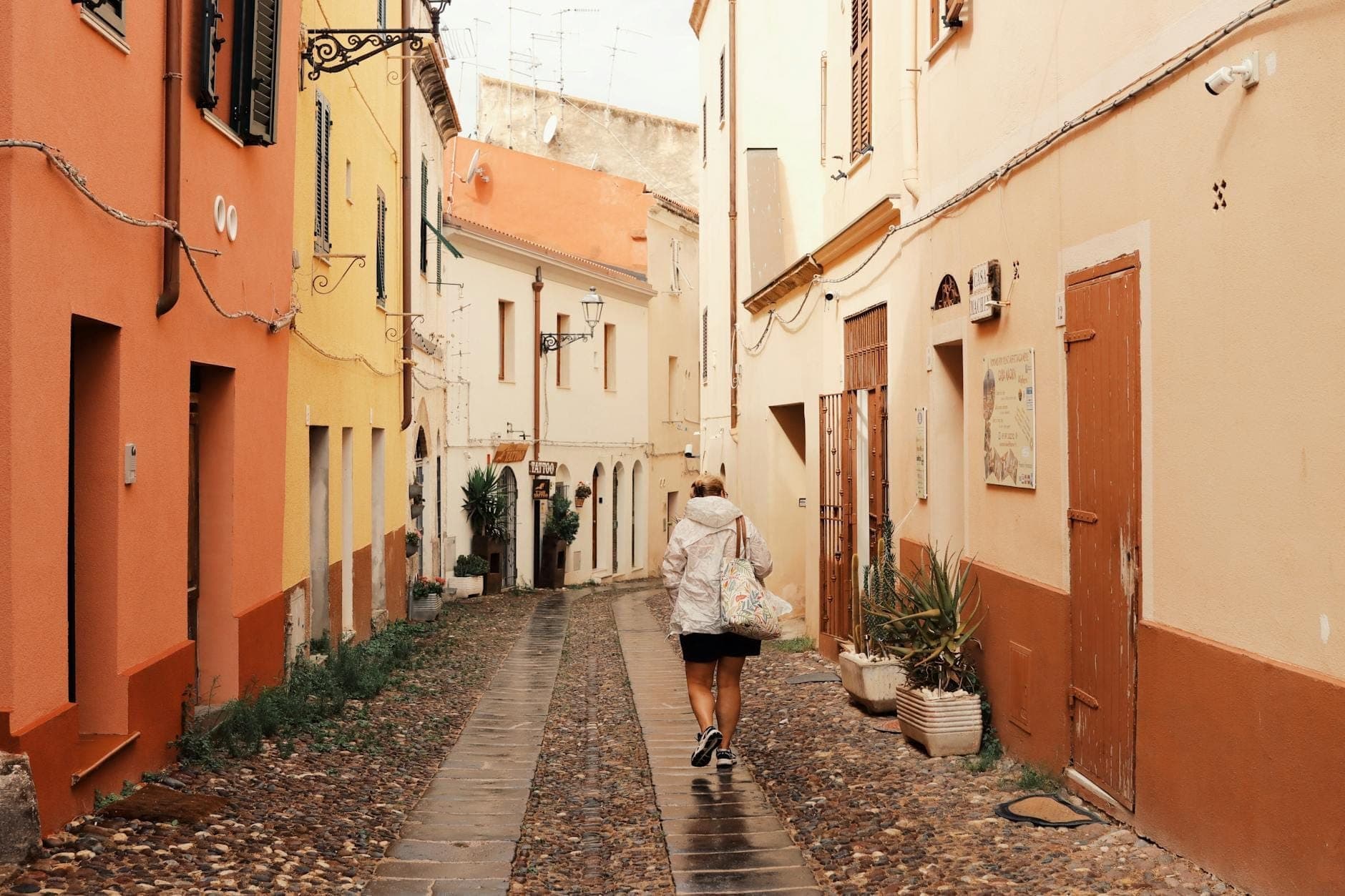Persona caminando por una encantadora calle estrecha adoquinada con coloridos edificios y plantas en macetas, alejándose de la costa de Cerdeña.