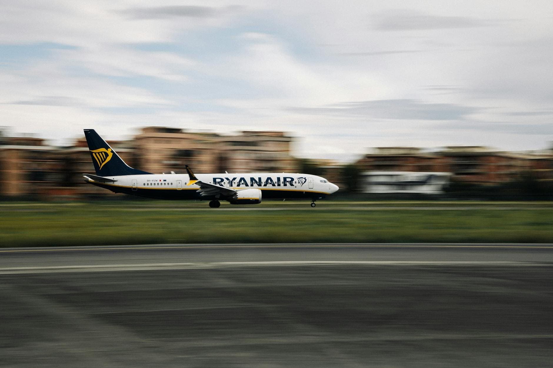 Un avión de Ryanair despegando o aterrizando en una pista con edificios borrosos al fondo.