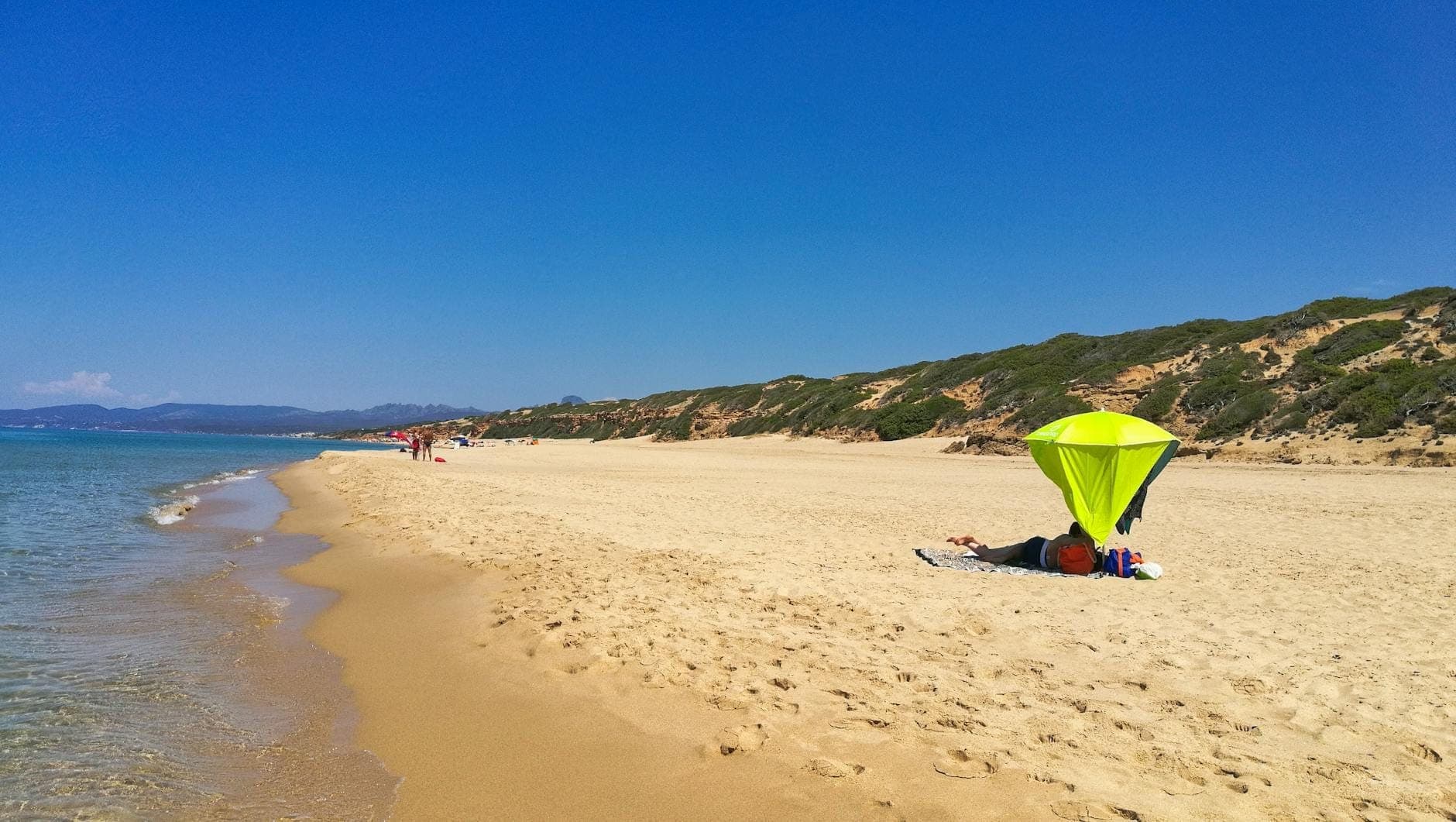 Breiter Sandstrand auf Sardinien an einem sonnigen Tag mit wenigen Menschen und einem grünen Sonnenschirm, der die geringe Besucherzahl und die Sommerhitze hervorhebt.
