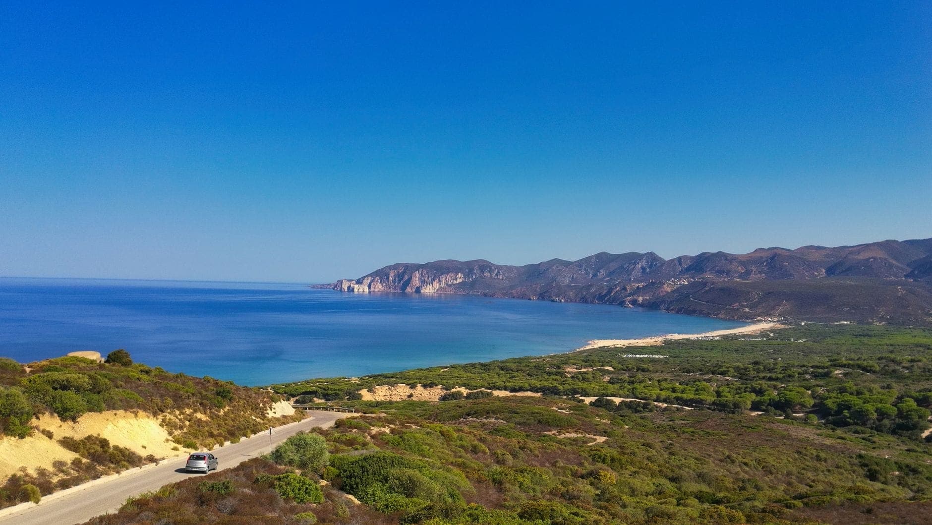 Una carretera costera sinuosa en Cerdeña con un coche circulando por los acantilados sobre el azul del Mediterráneo y montañas escarpadas bajo un cielo despejado.