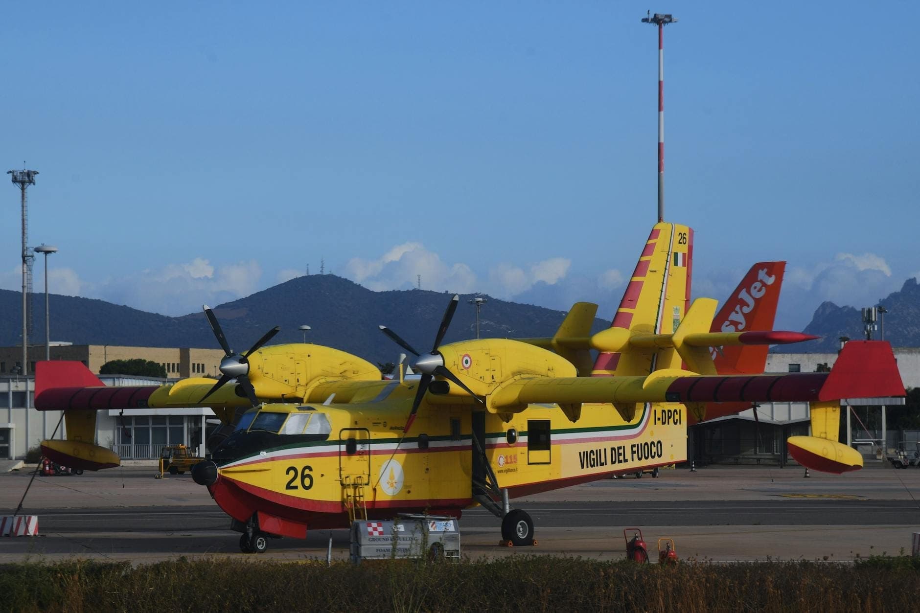 Ein gelbes Löschflugzeug auf dem Rollfeld eines Flughafens, mit Terminalgebäuden und Bergen im Hintergrund unter einem blauen Himmel.