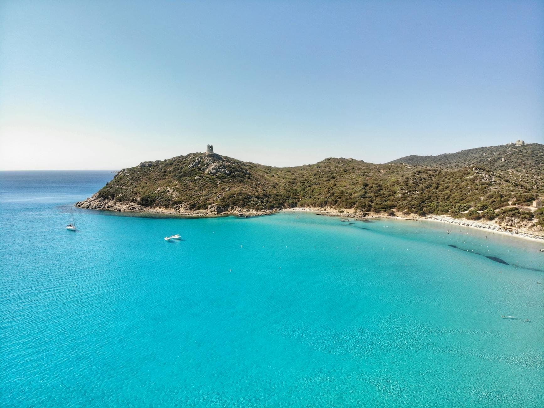 Vista aérea de una bahía turquesa con agua cristalina, botes y una torre de vigilancia costera en el sureste de Cerdeña cerca de Villasimius.