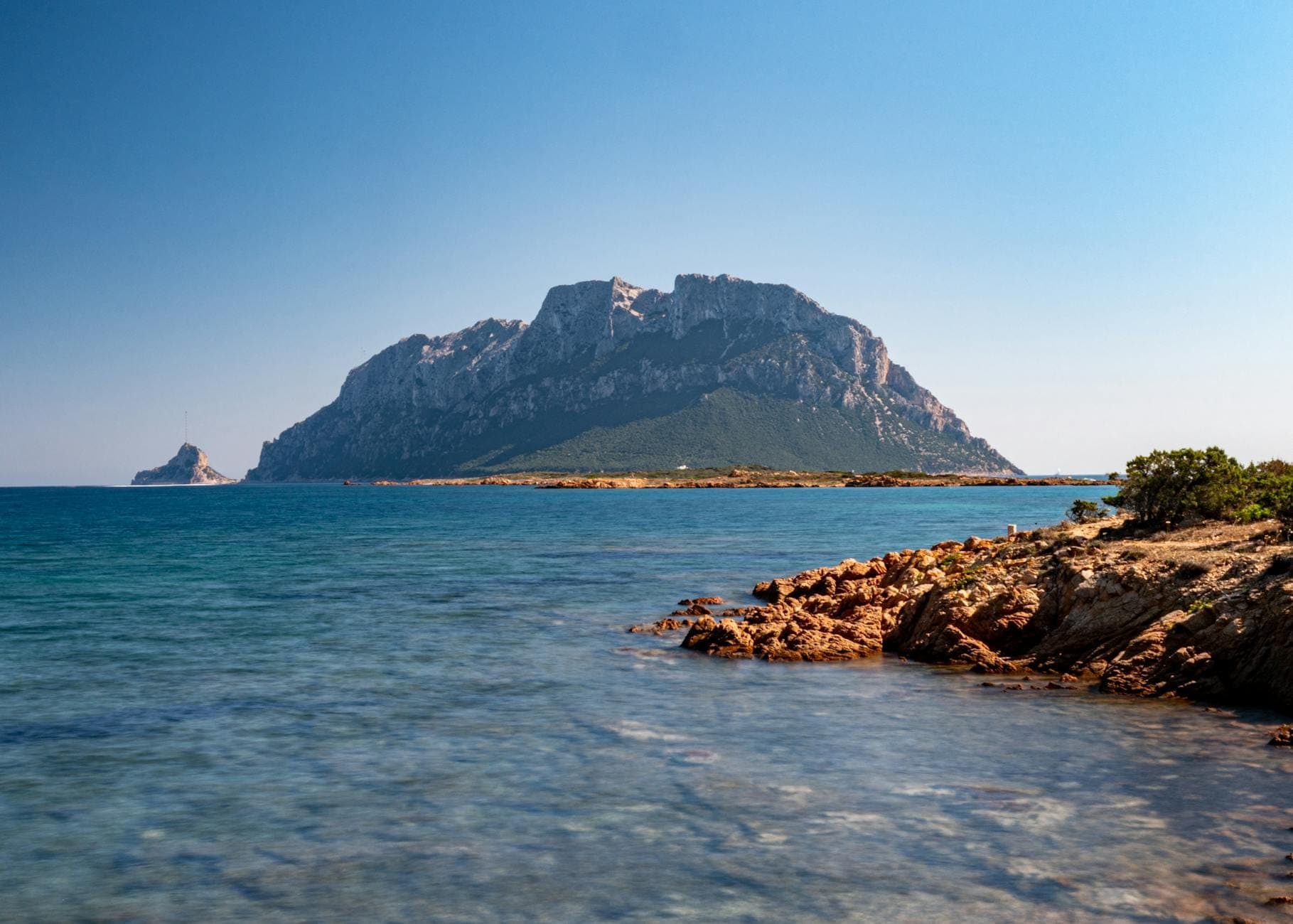 Vista despejada de la Isola Tavolara emergiendo del mar turquesa, vista desde una costa rocosa del noreste de Cerdeña bajo un cielo azul brillante.