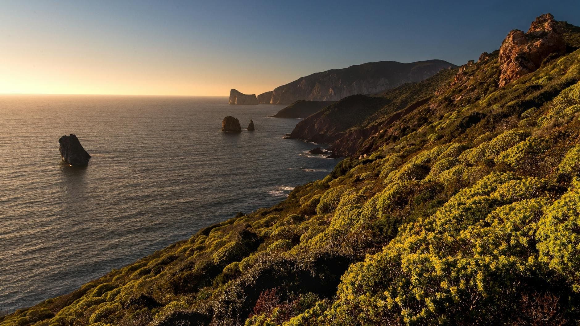 Espectacular costa rocosa del suroeste de Cerdeña al atardecer, con acantilados escarpados, islotes rocosos y exuberante matorral mediterráneo con vistas al mar azul.