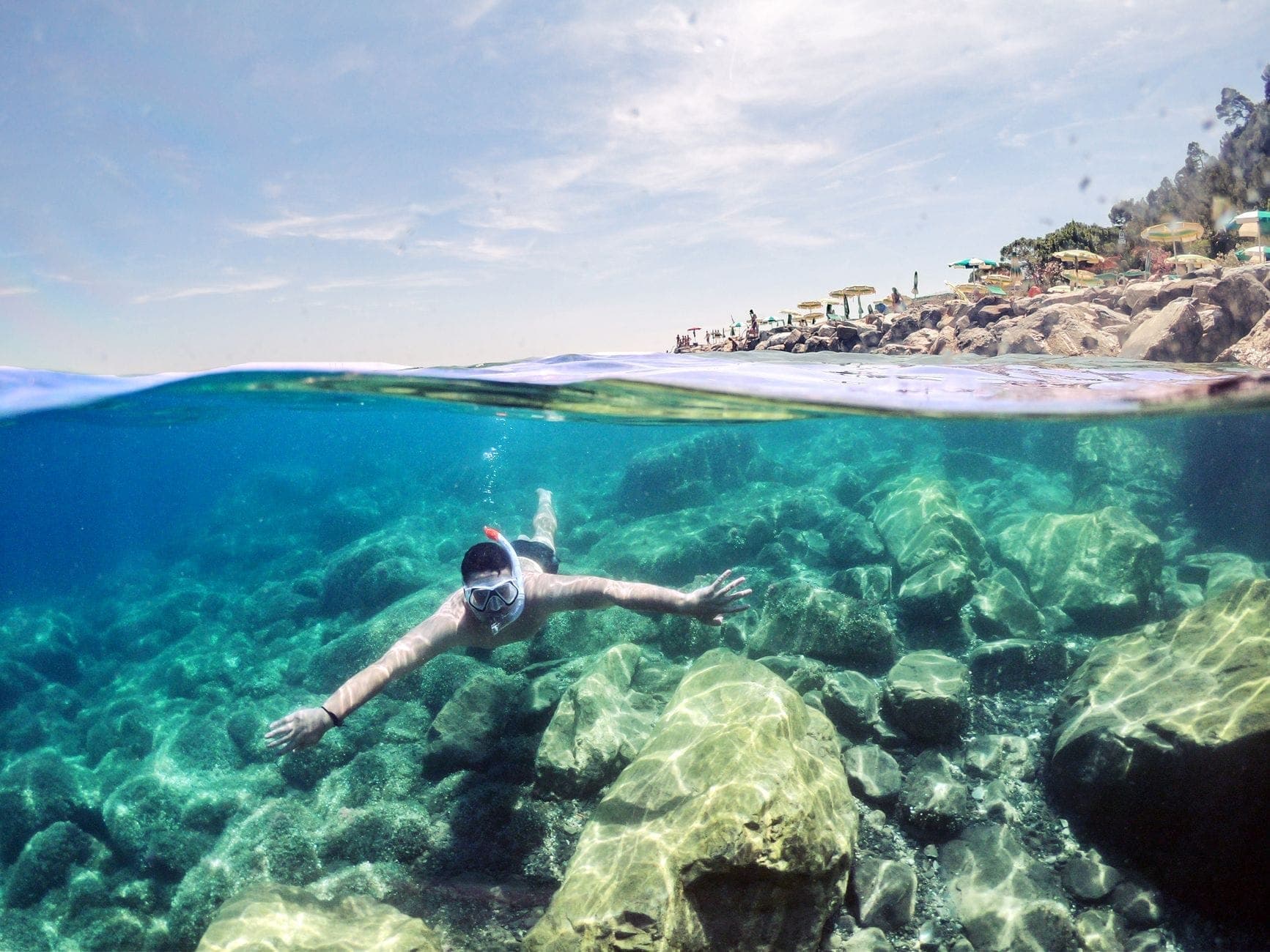 Buceador con tubo en aguas azules y cristalinas sobre un fondo rocoso cerca de una costa mediterránea, con una playa iluminada por el sol y personas al fondo.