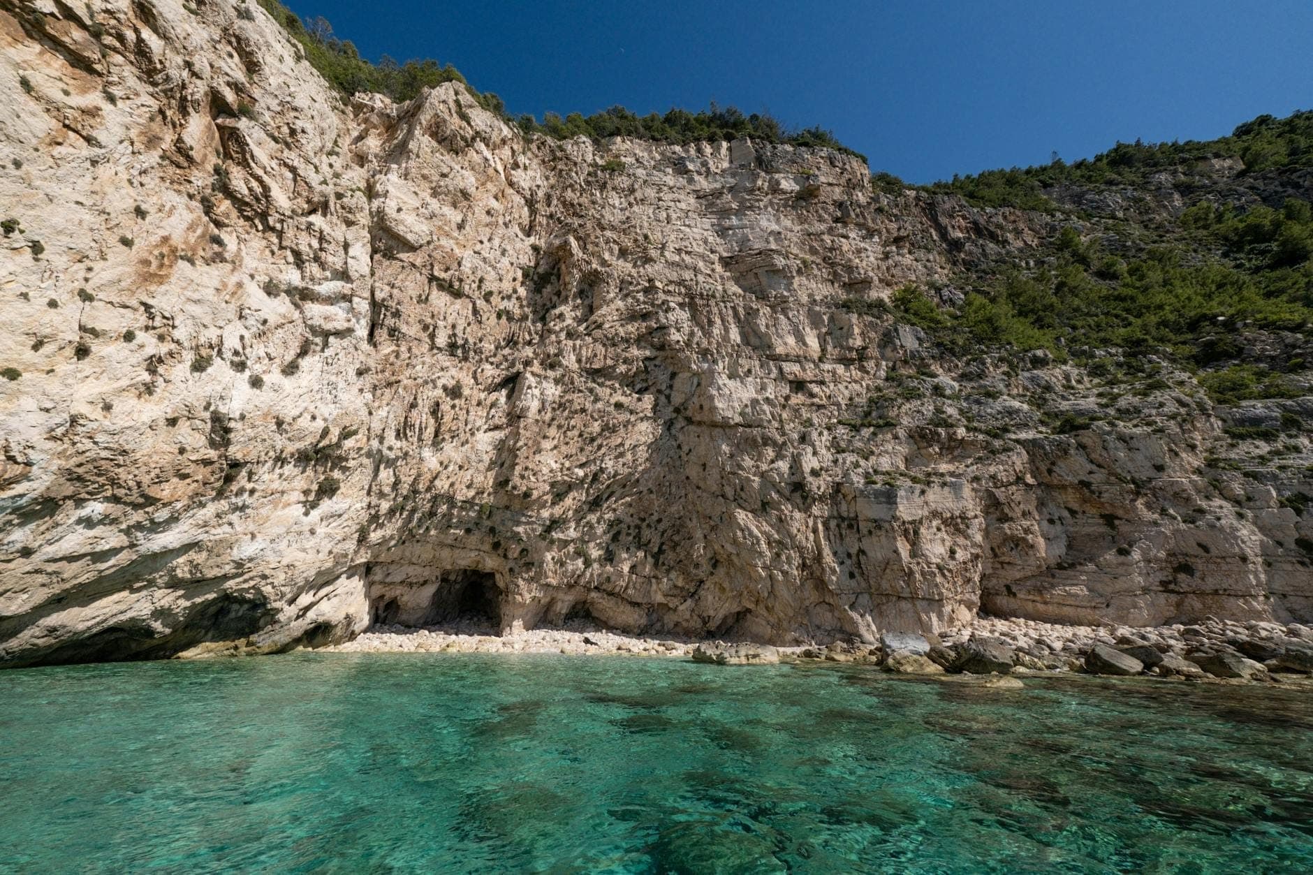 Agua turquesa con escarpados acantilados de piedra caliza y pequeñas cuevas marinas a lo largo de la costa de Cerdeña bajo un cielo azul despejado.