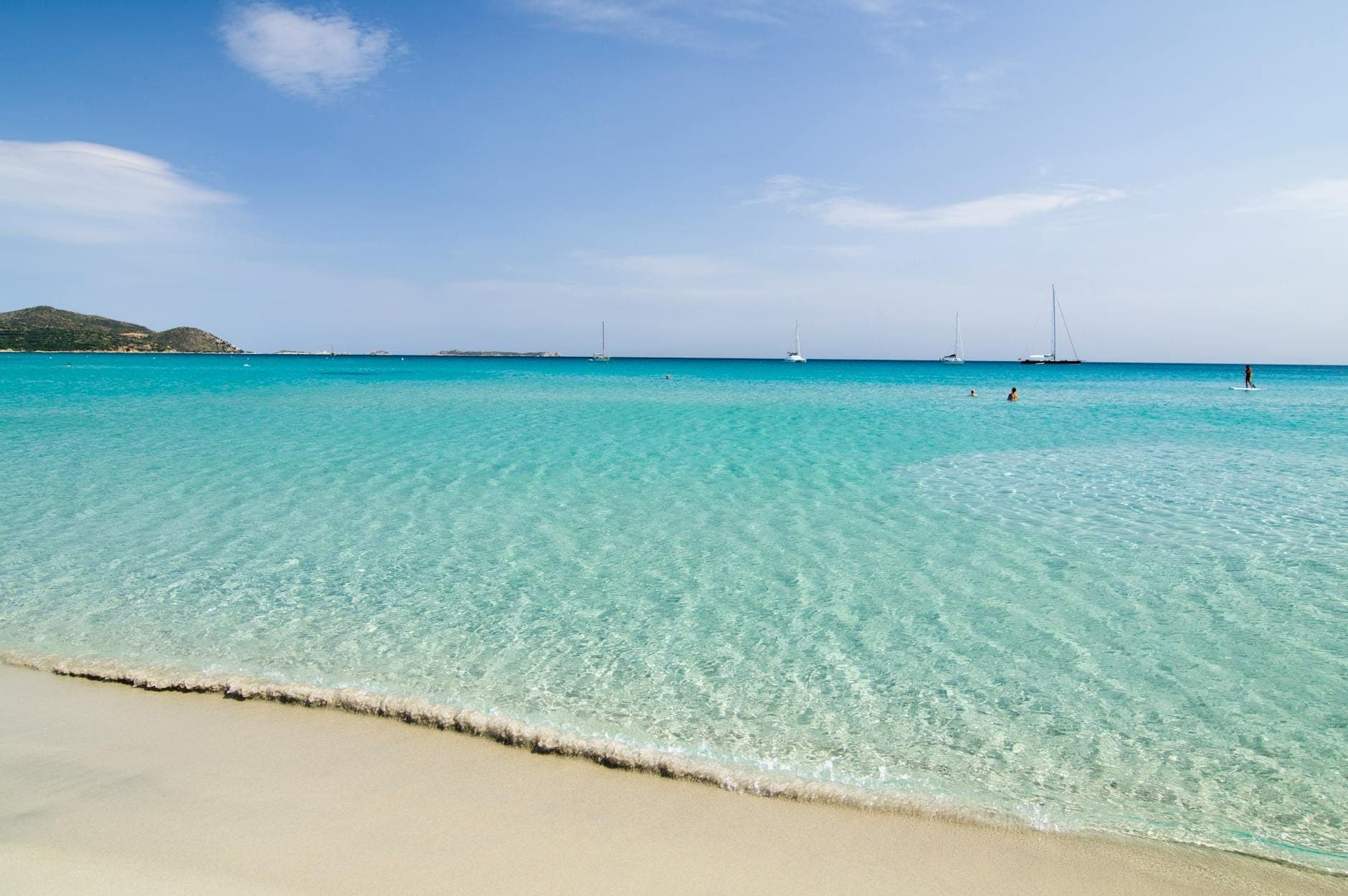 Kristallklares flaches Wasser mit einem ruhigen Sandstrand, türkisfarbenem Meer und einigen vor Anker liegenden Segelbooten im Süden Sardiniens.