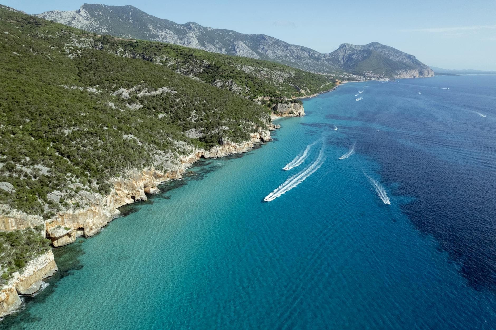 Vista aérea de la accidentada costa de Cerdeña con agua turquesa, acantilados de piedra caliza, barcos y paisaje montañoso al fondo.