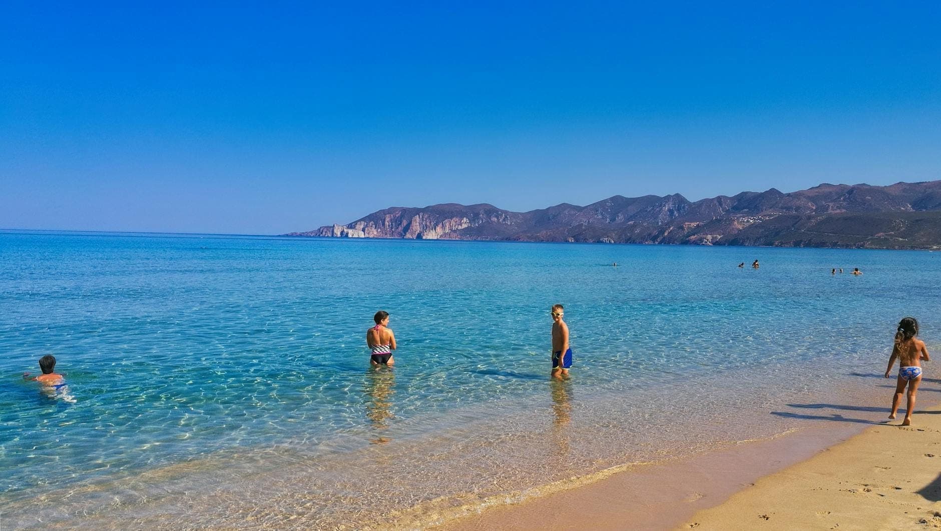 Niños y familias disfrutando de aguas turquesas poco profundas y una playa de arena con montañas al fondo en un día soleado en Cerdeña.