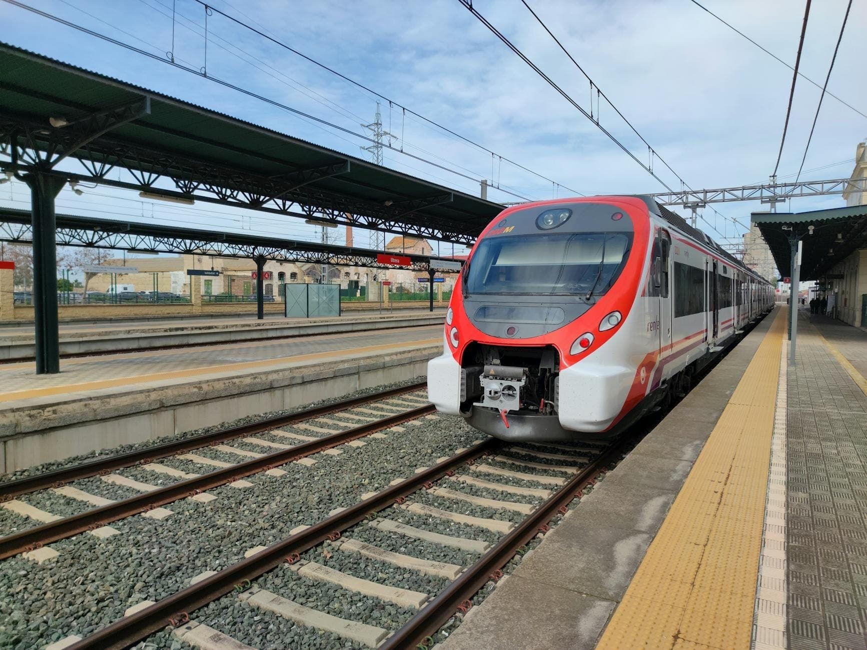 Tren moderno de Trenitalia en rojo y blanco en el andén de una estación bajo un cielo azul, mostrando el transporte ferroviario regional de Cerdeña.