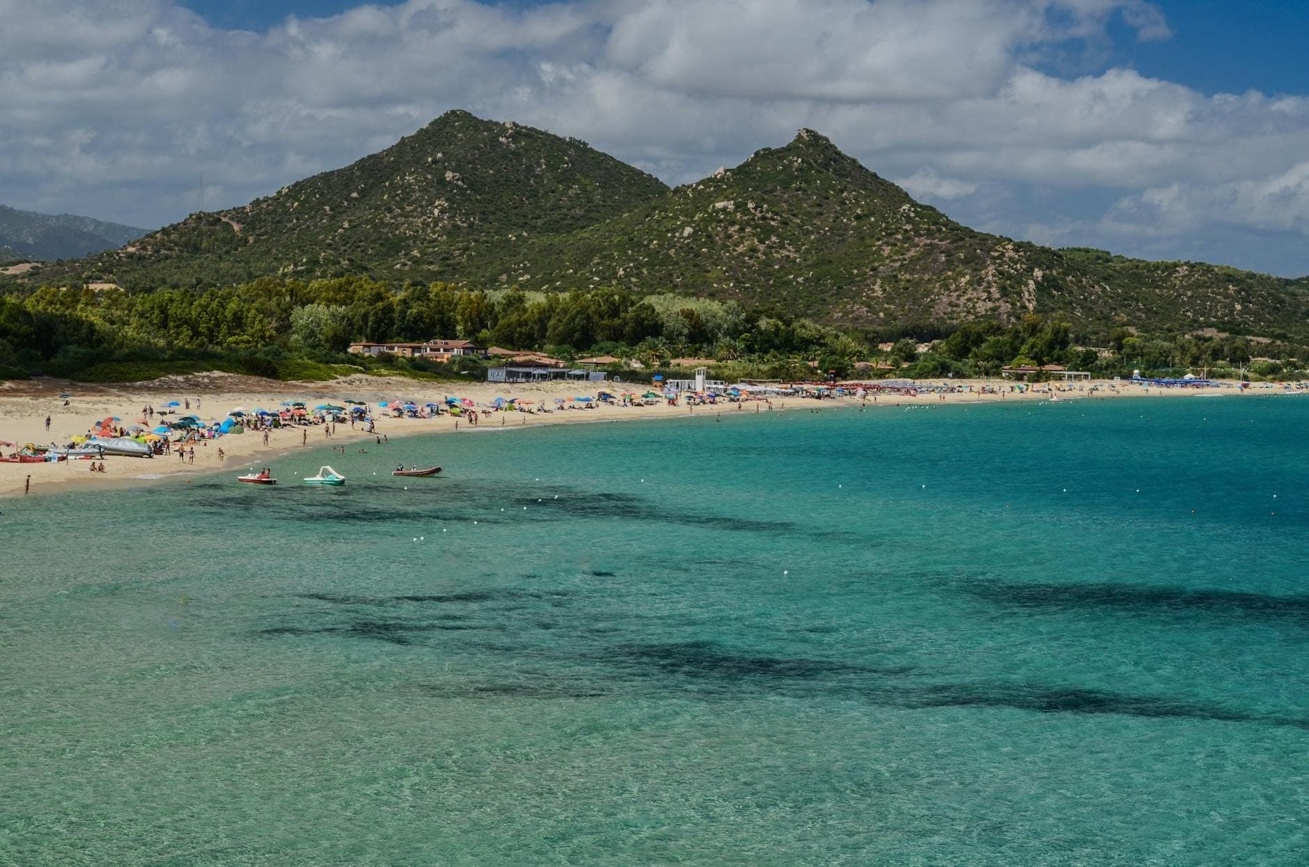 Una amplia playa de Cerdeña con agua turquesa, olas suaves, sombrillas, familias y colinas verdes a lo lejos bajo un cielo parcialmente nublado.