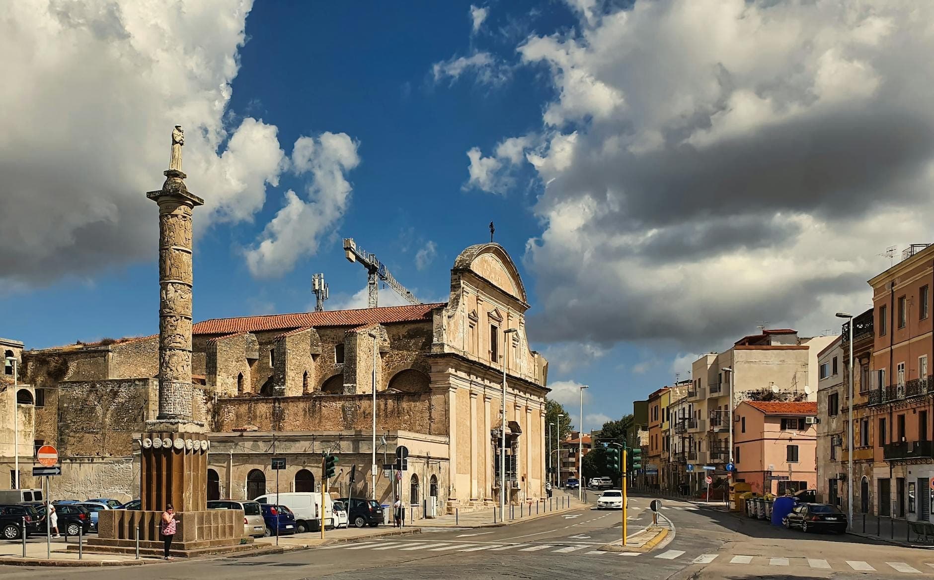 Una amplia plaza de Sassari, Cerdeña, rodeada de edificios históricos y una iglesia, con una columna destacada y nubes dramáticas sobre ella.
