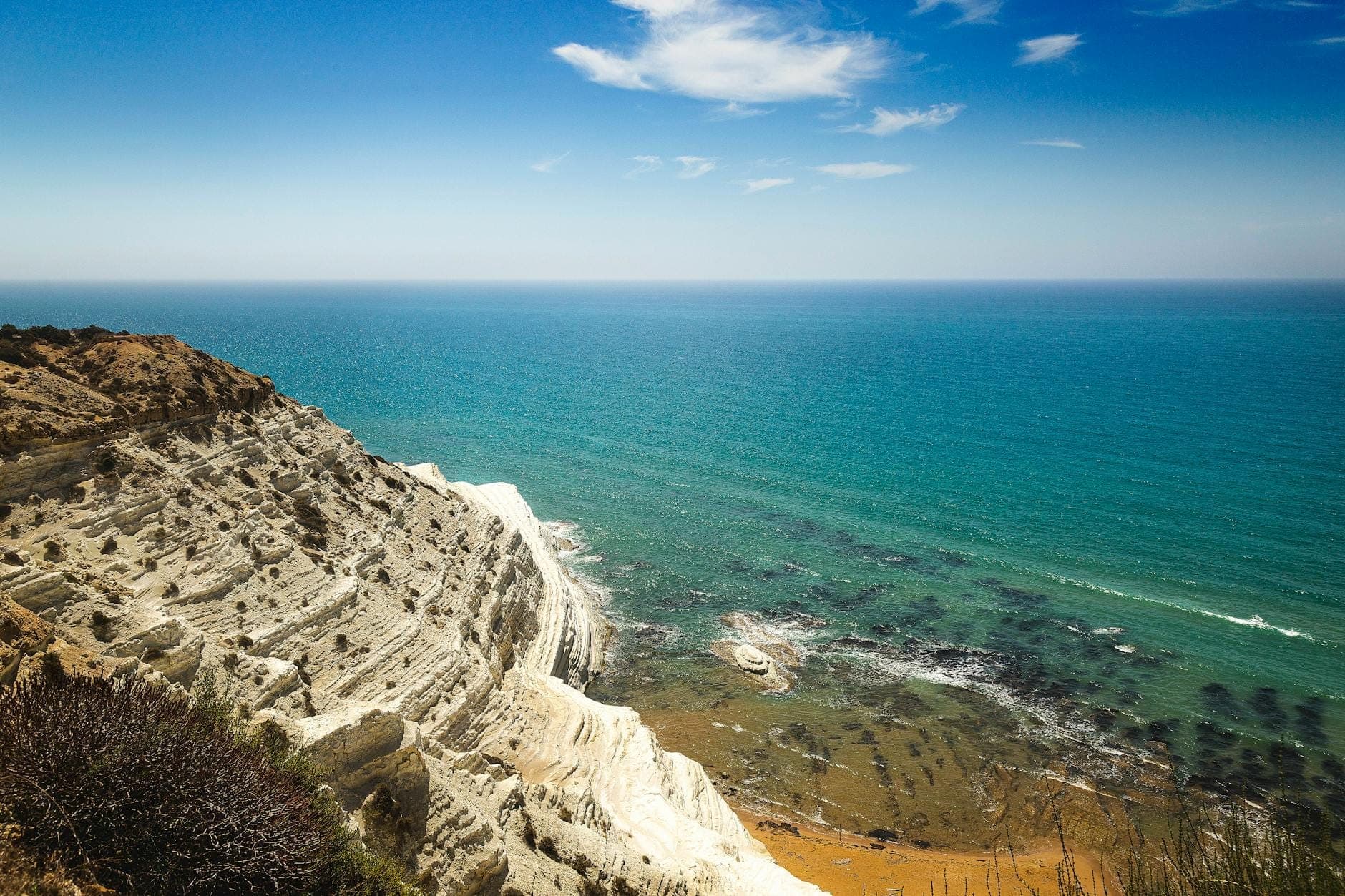 Pemandangan luas tebing marl putih ikonik Scala dei Turchi yang bertingkat menuju Laut Mediterania berwarna toska di bawah langit biru cerah.