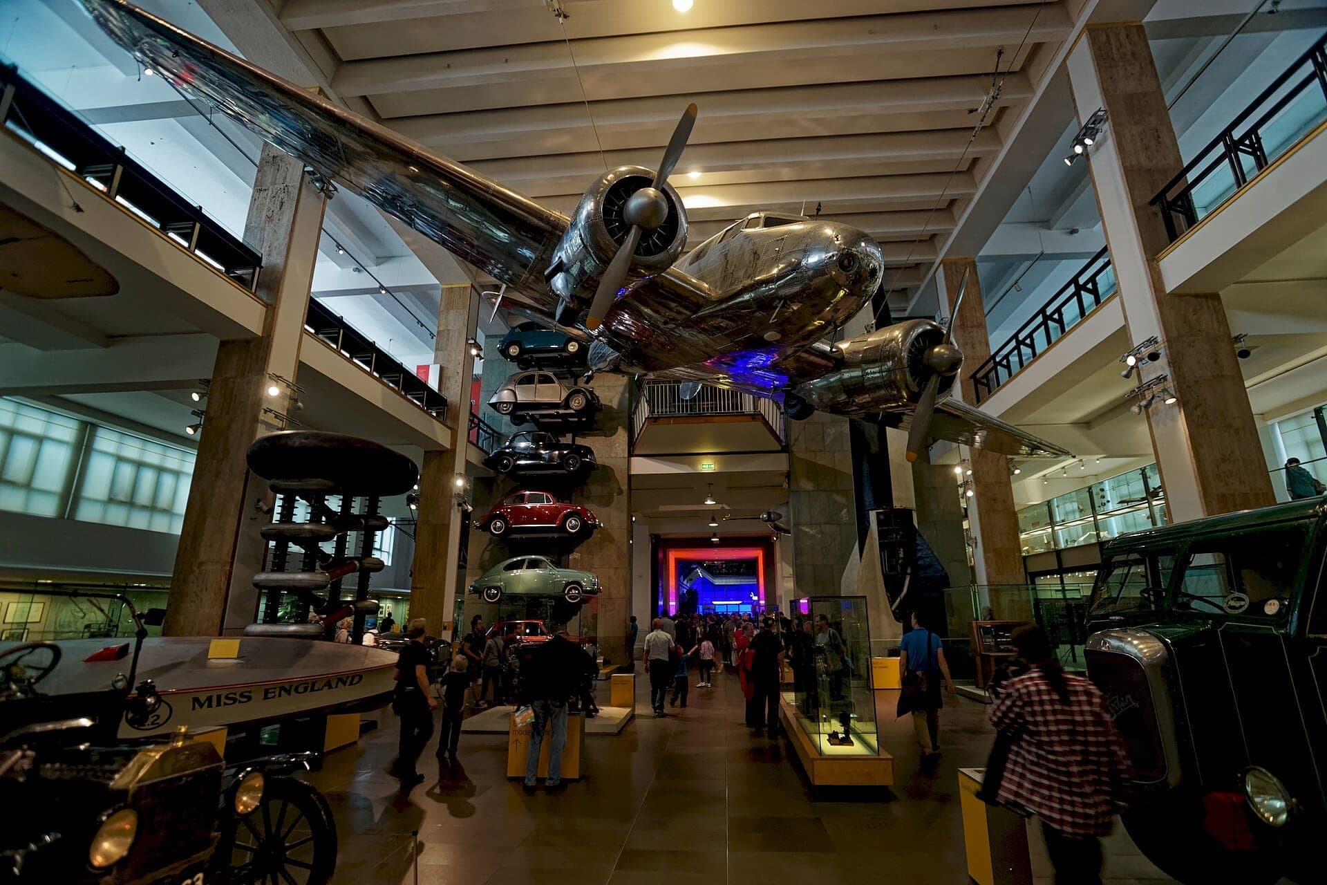 Im Inneren des Science Museum London schlendern Besucher unter einem großen hängenden Flugzeug, umgeben von Oldtimern und historischen Fahrzeugen in einer geräumigen Halle.