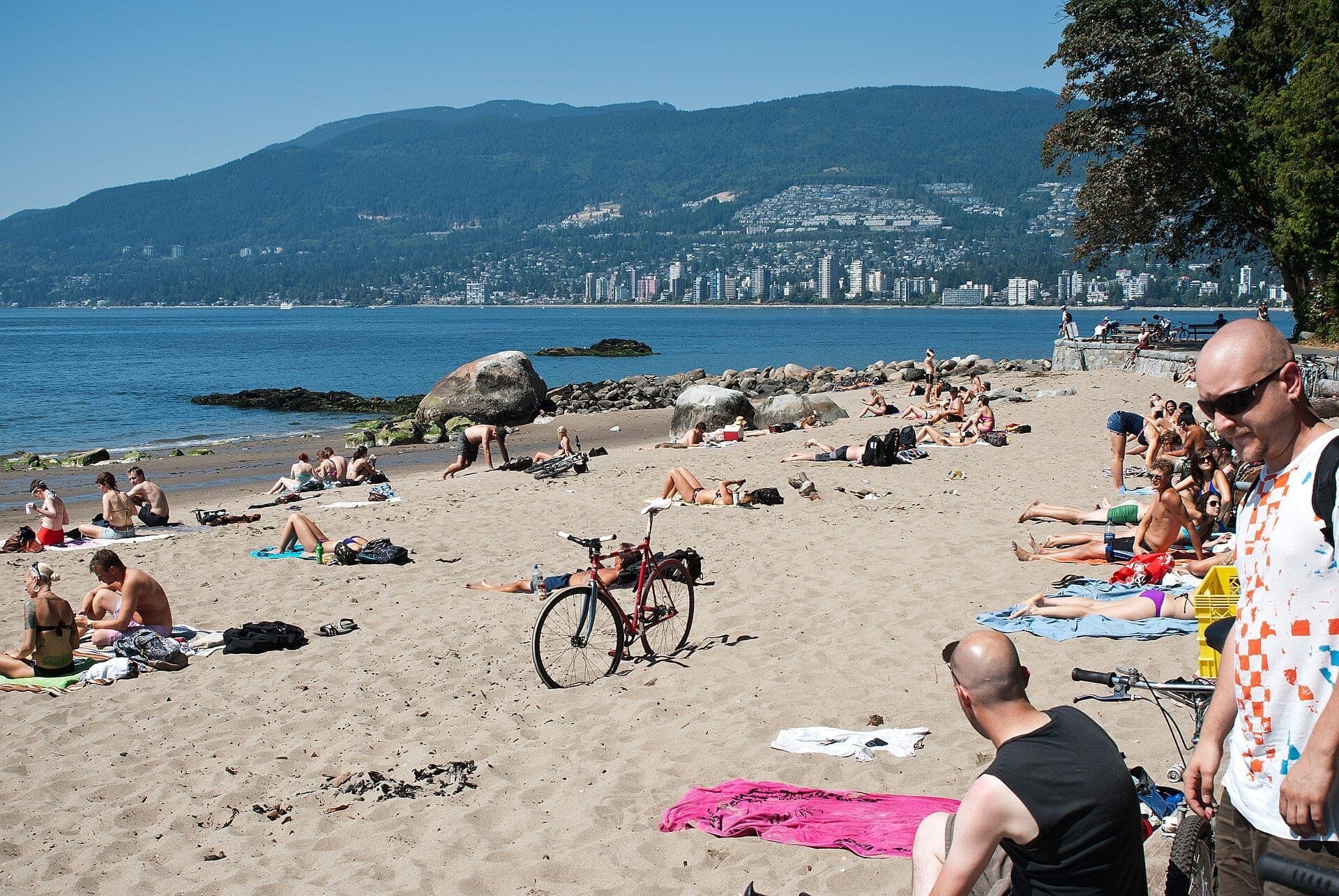 นักอาบแดดพักผ่อนบนชายหาดทรายของ Second Beach ใน Stanley Park พร้อมวิวภูเขาและเมืองฝั่งตรงข้าม English Bay ใต้ท้องฟ้าสีฟ้าแจ่มใส