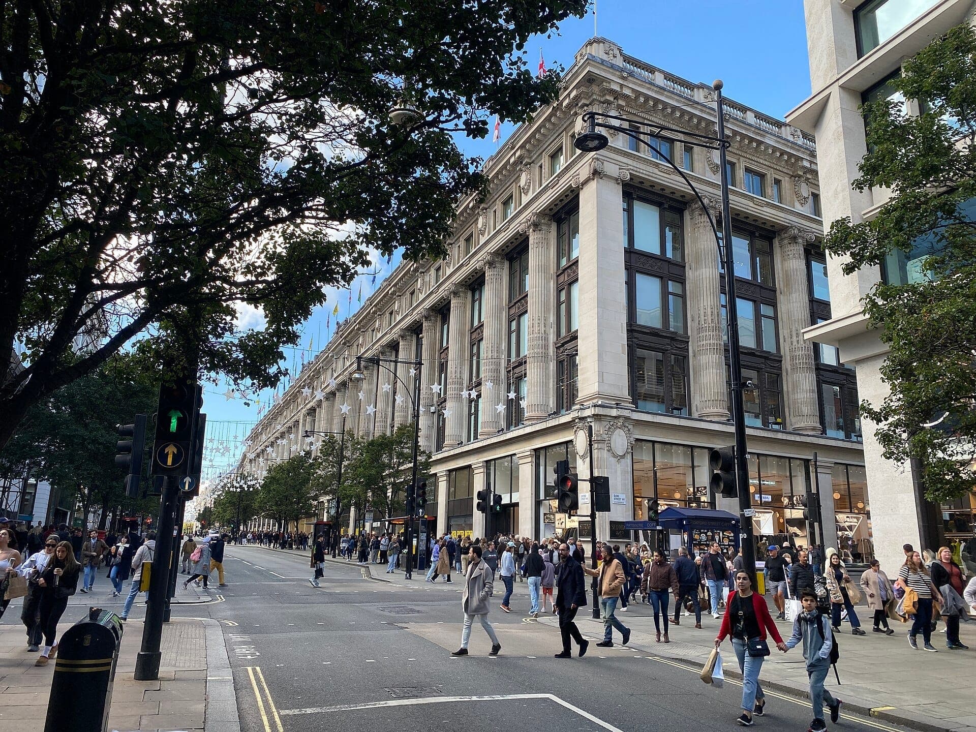 La grande façade de Selfridges Londres sur une Oxford Street animée, avec des foules de clients traversant sous un ciel bleu clair.