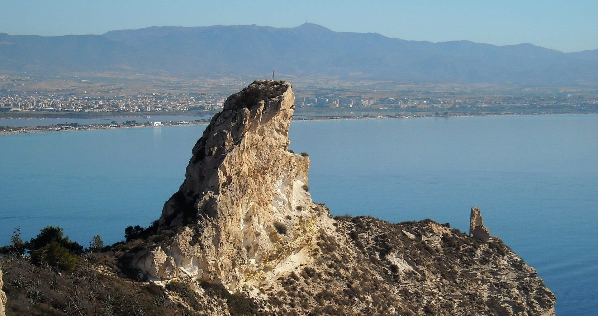 Prominente afloramiento de piedra caliza de la Sella del Diavolo que se eleva sobre la costa azul, con la ciudad de Cagliari y las montañas visibles al fondo.