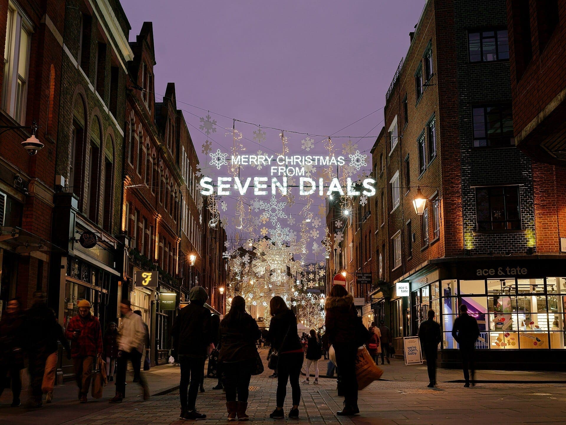 Evening view of Seven Dials in London with festive Christmas lights, snowflake decorations, and people walking along historic brick buildings.