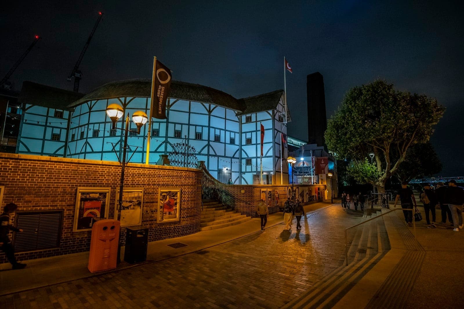 Exterior view of Shakespeare's Globe Theatre in London at night, illuminated with blue lighting, with people walking along the lit pathway in front.
