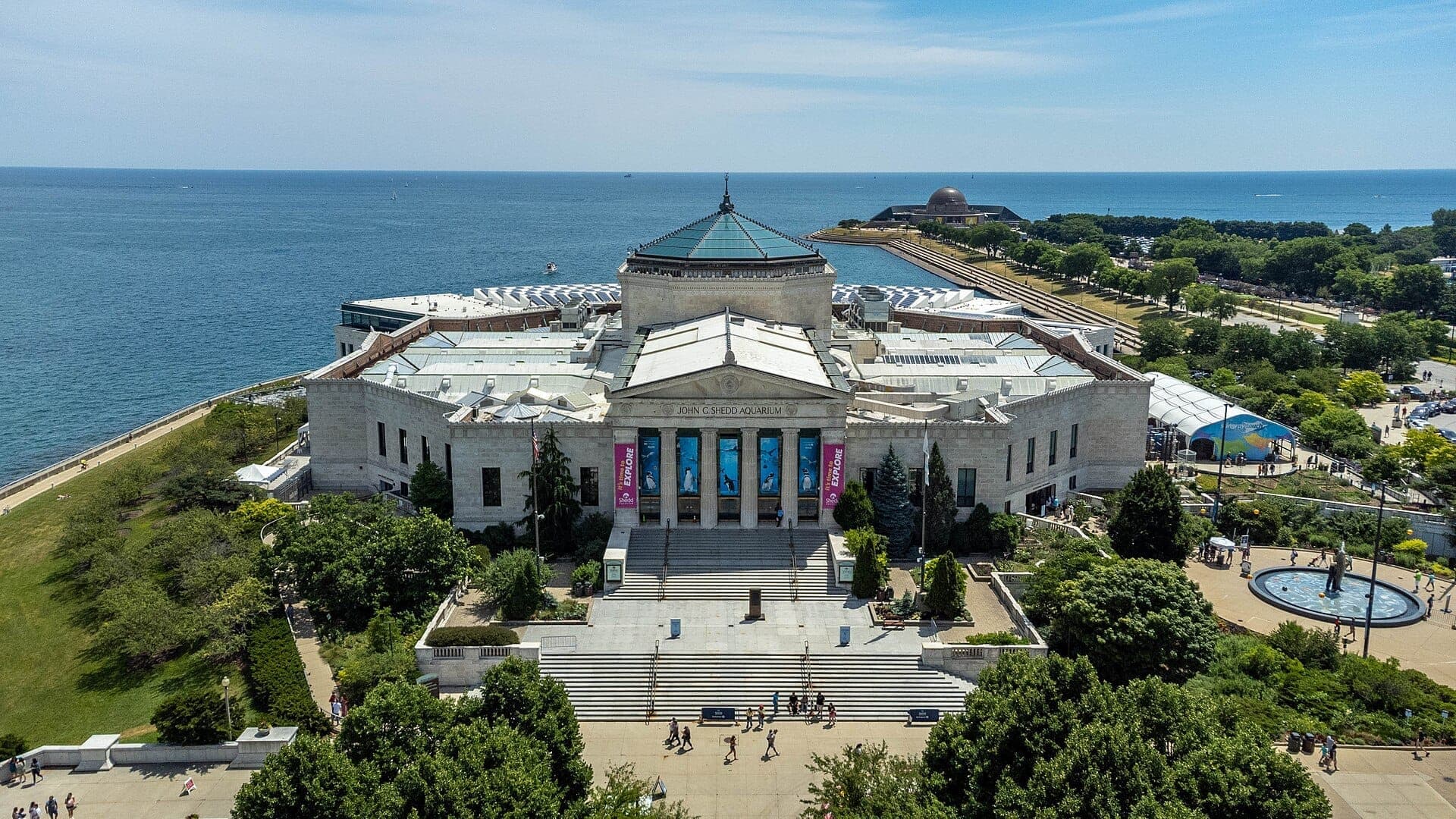 Aerial view of Shedd Aquarium in Chicago, featuring its grand entrance, surrounding greenery, and Lake Michigan in the background on a sunny day.