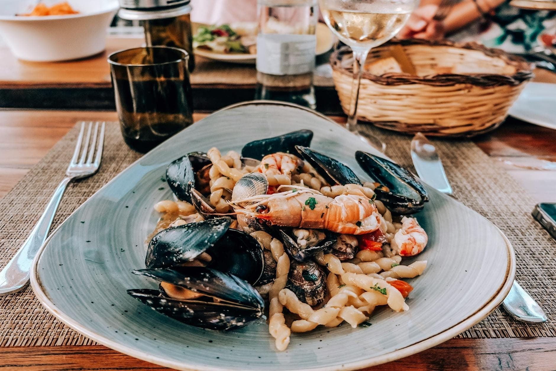 Pasta seafood dengan kerang dan udang tersaji indah di atas meja bersama gelas anggur, mencerminkan suasana santai restoran khas Sisilia.