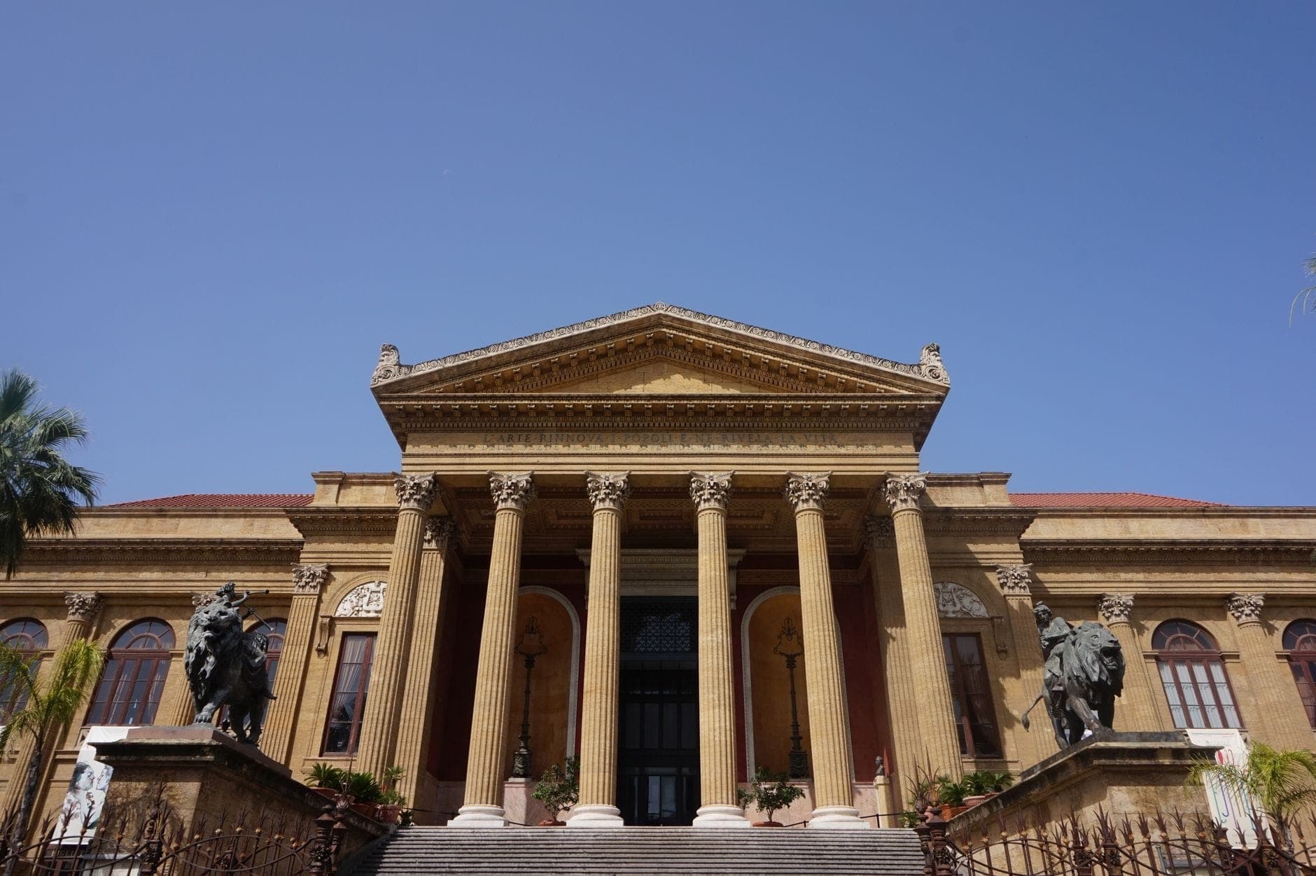 Tampak depan Teatro Massimo di Palermo dengan kolom-kolom megah dan patung-patung, di bawah langit biru yang cerah.