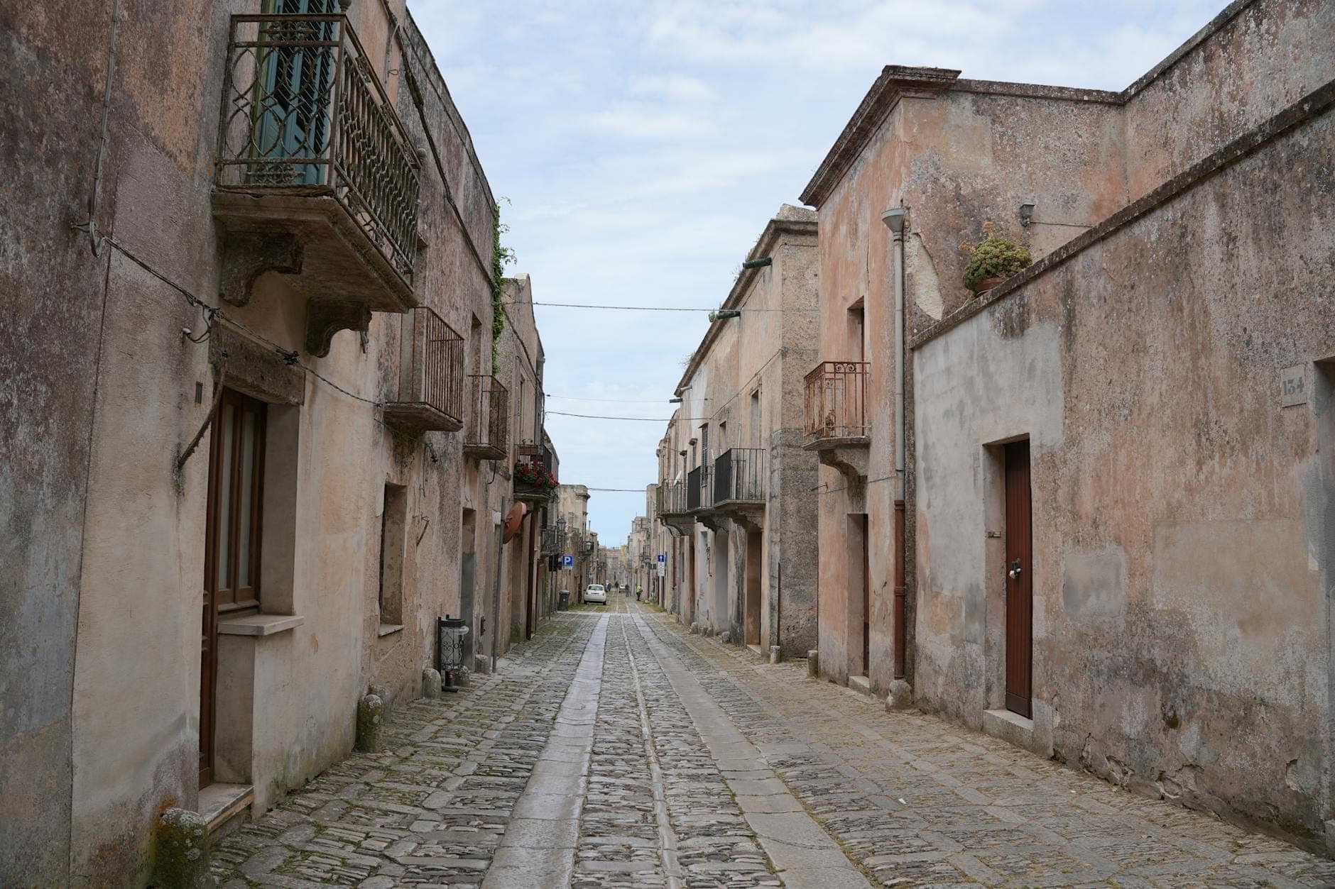 Jalan berbatu lebar yang diapit bangunan-bangunan batu tua di sebuah desa Sisilia yang tenang, membangkitkan suasana klasik film-film The Godfather.