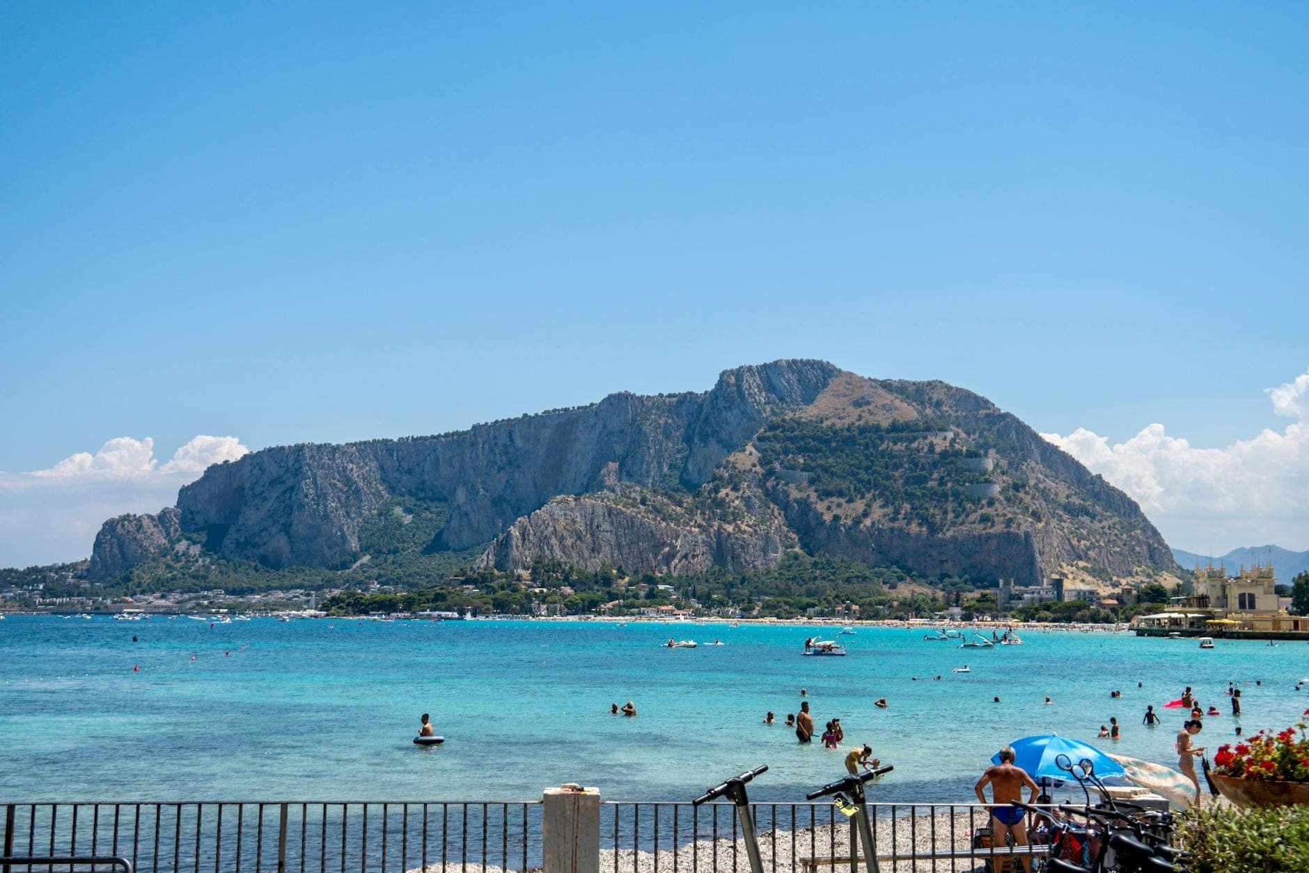 Pemandangan jelas Pantai Mondello di Sisilia dengan perenang di laut, air biru kehijauan yang cerah, pegunungan di latar belakang, dan hari yang cerah.
