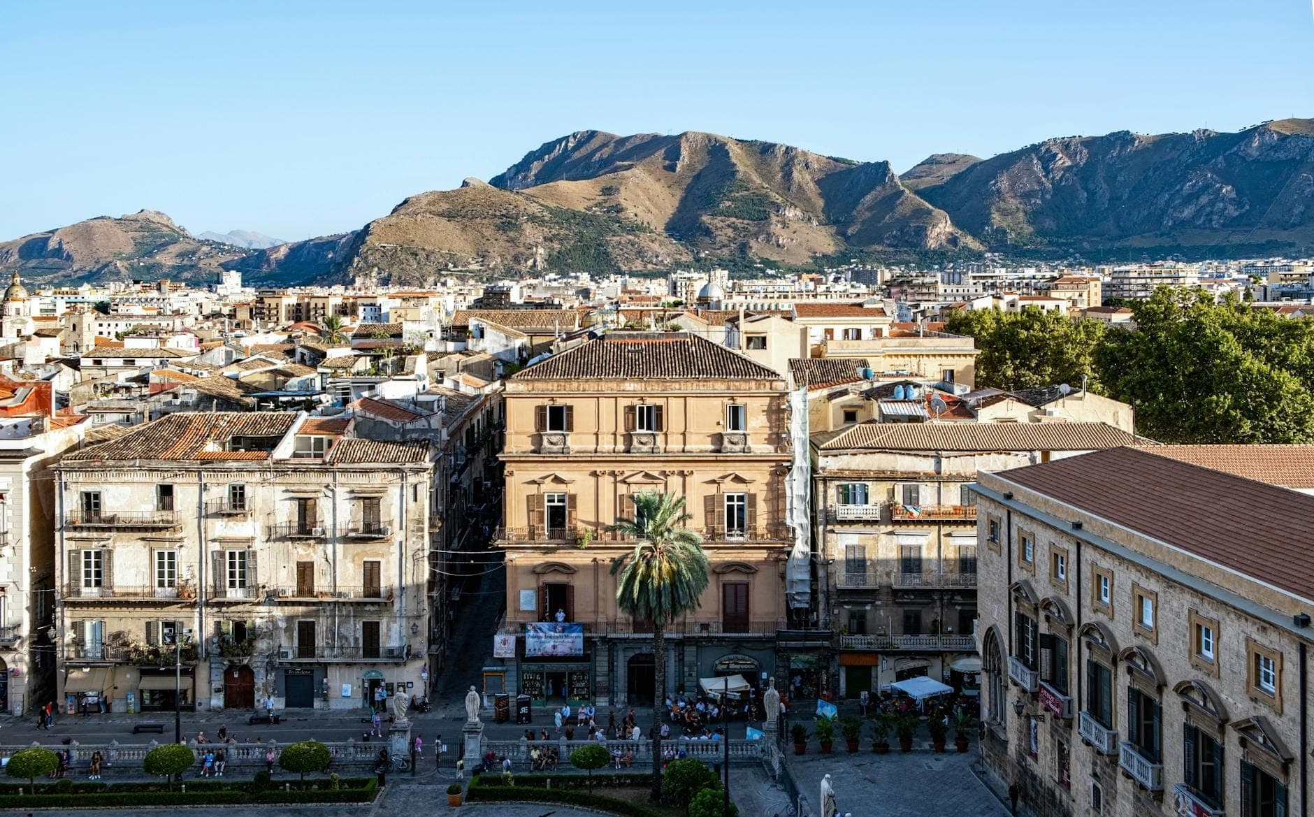 Pemandangan panoramik Palermo, Sisilia yang diterangi cahaya matahari, dengan bangunan bersejarah, pegunungan di latar belakang, dan langit biru cerah yang khas musim panas Mediterania yang panas dan kering.