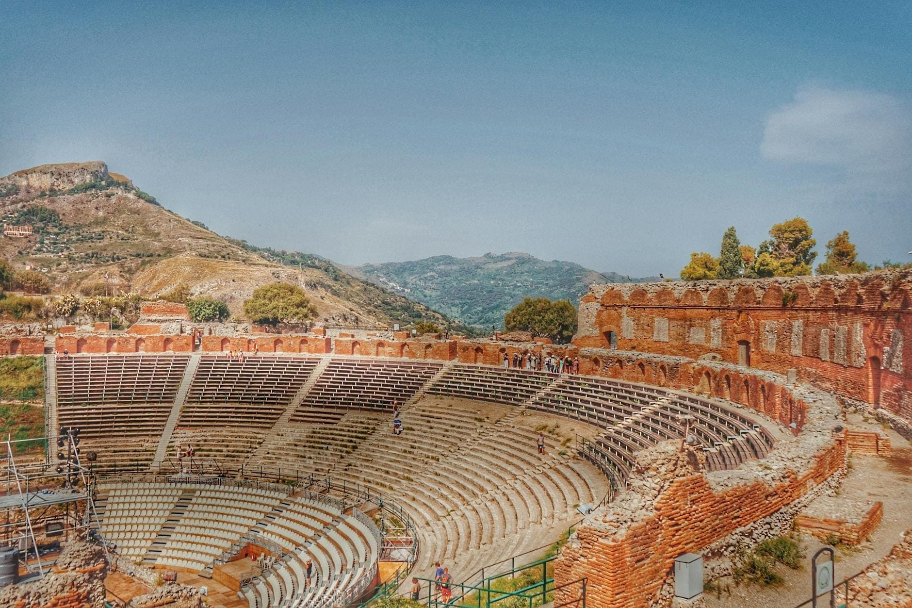 Pemandangan luas Teatro Greco di Taormina yang bersejarah dengan bukit-bukit di sekitarnya dan langit biru, venue ikonik untuk festival dan acara musim panas di Sisilia.