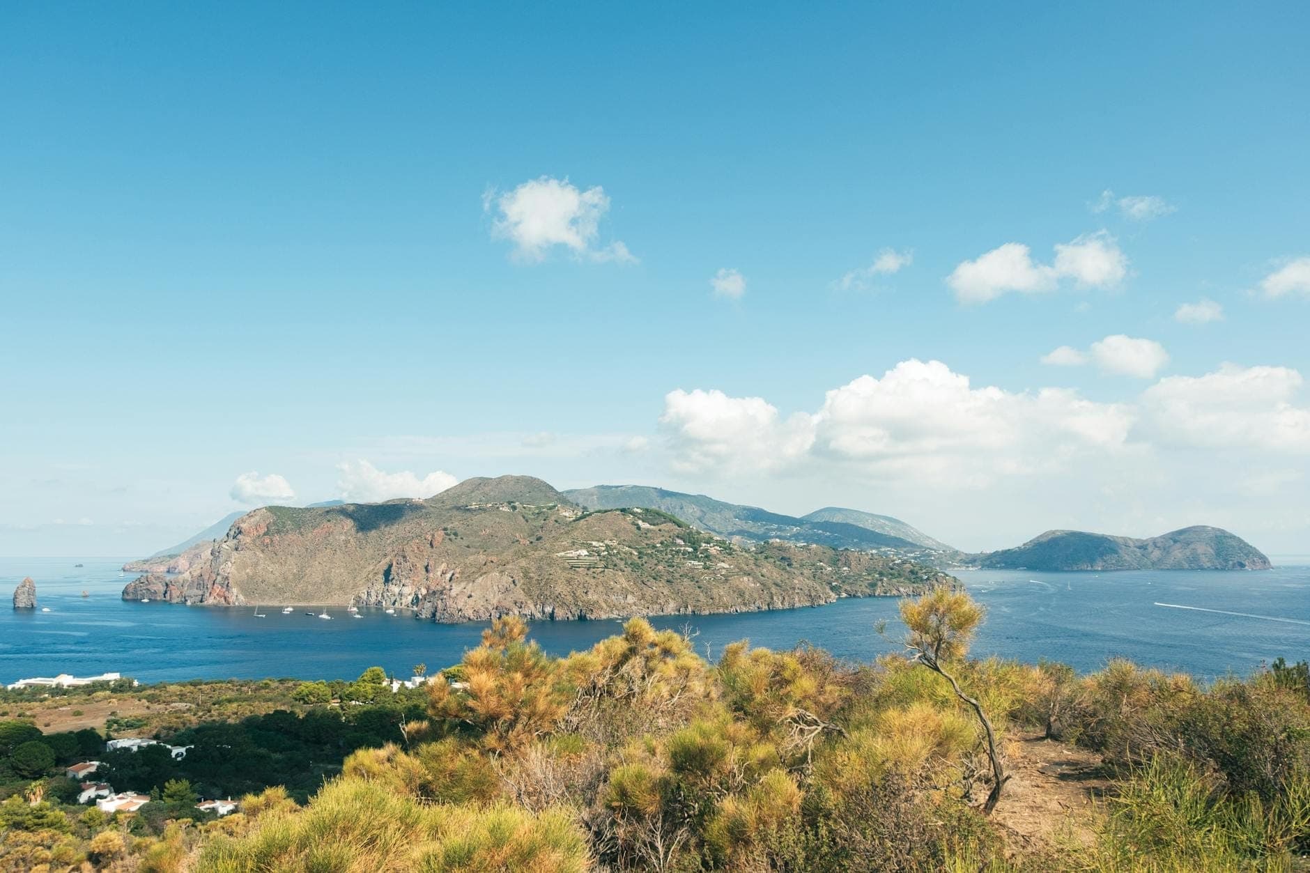 Pemandangan panoramik pulau vulkanik Aeolian yang dikelilingi laut biru tua, pulau-pulau kecil di kejauhan, awan tipis, dan vegetasi Mediterania di latar depan.