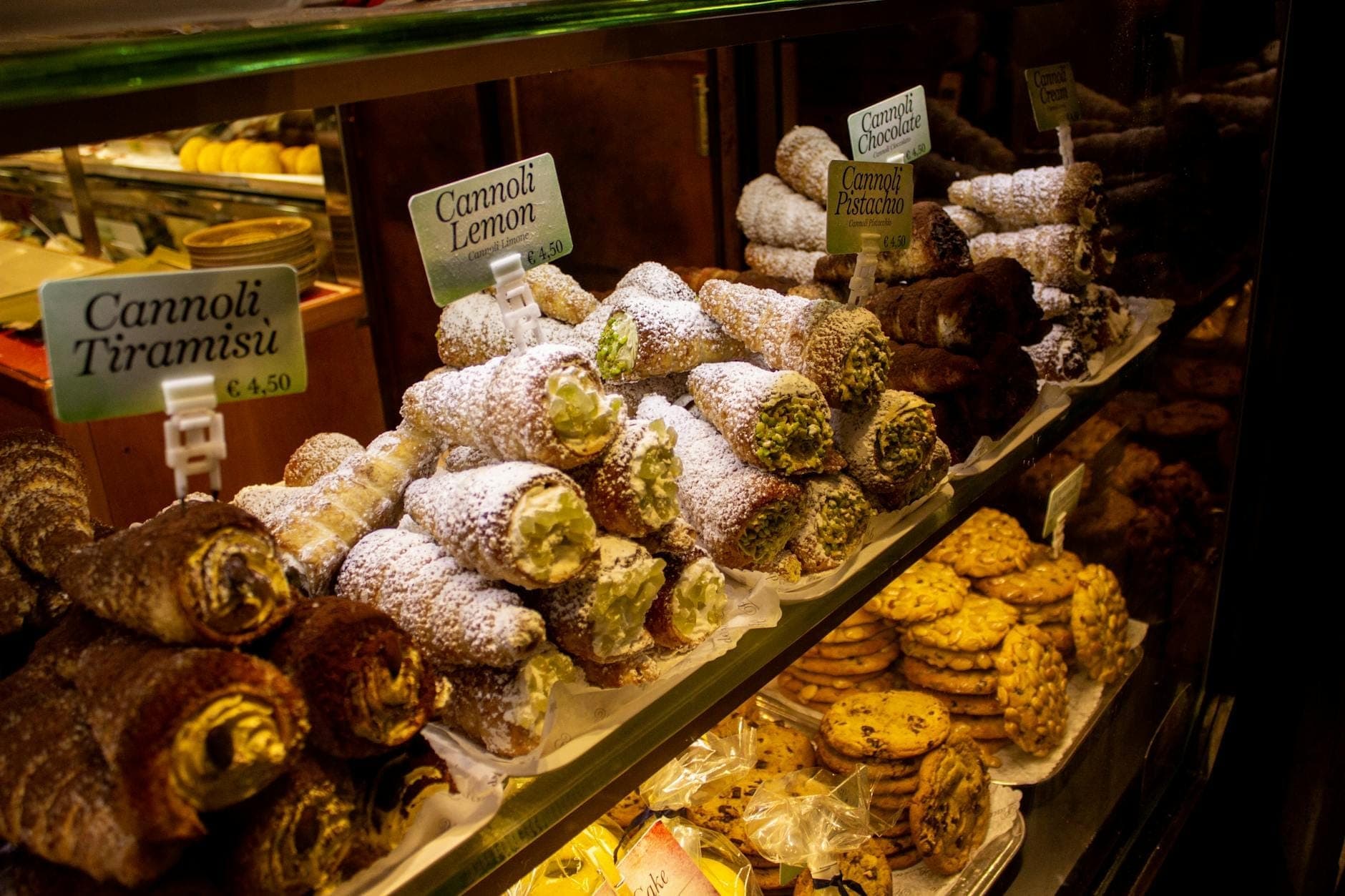 Lemari kaca berisi berbagai rasa cannoli yang ditaburi gula halus di sebuah toko kue Sisilia.