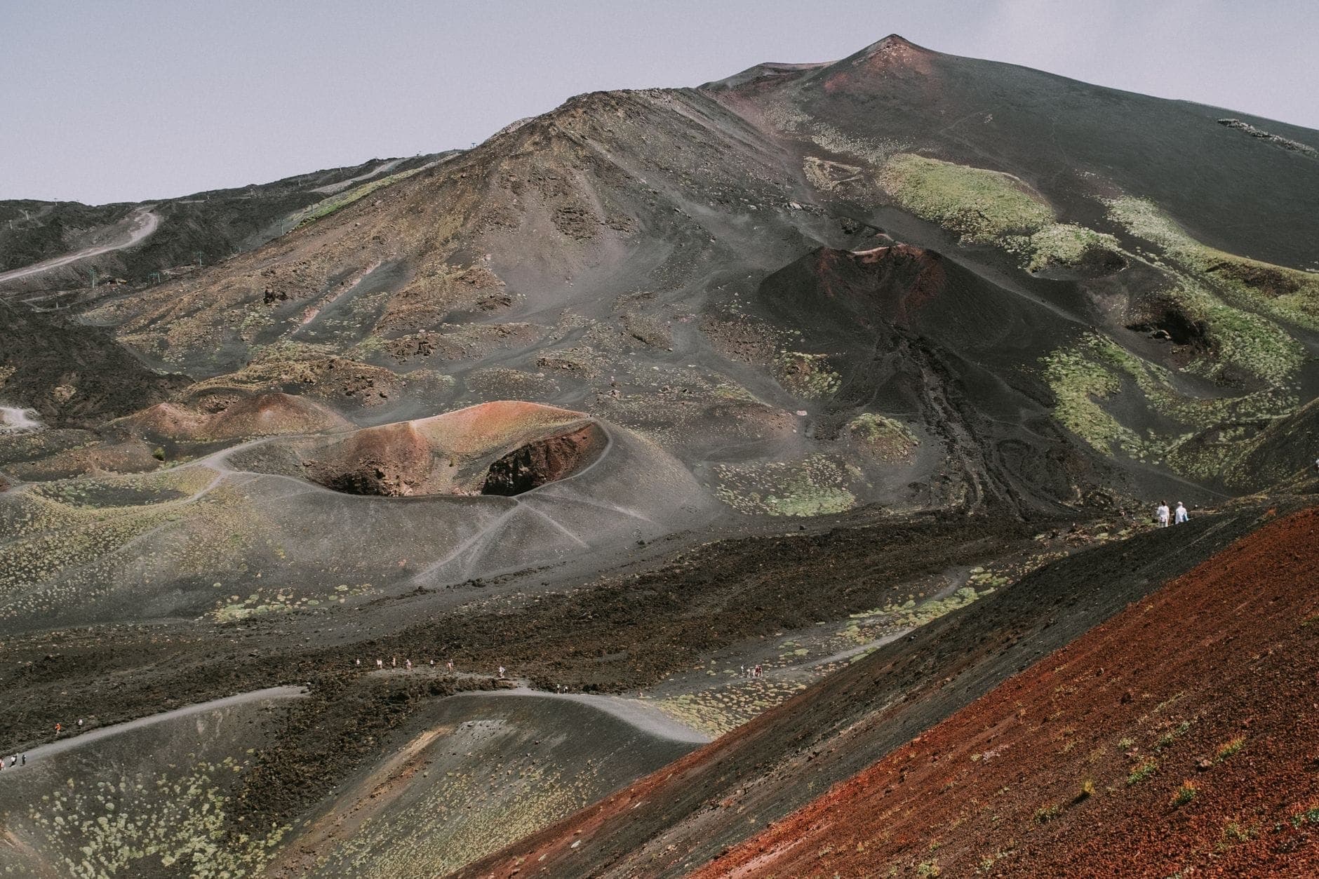 Pemandangan kawah vulkanik Gunung Etna dari dekat dengan bebatuan berwarna-warni dan jalur pendakian yang terlihat di bawah langit berawan.