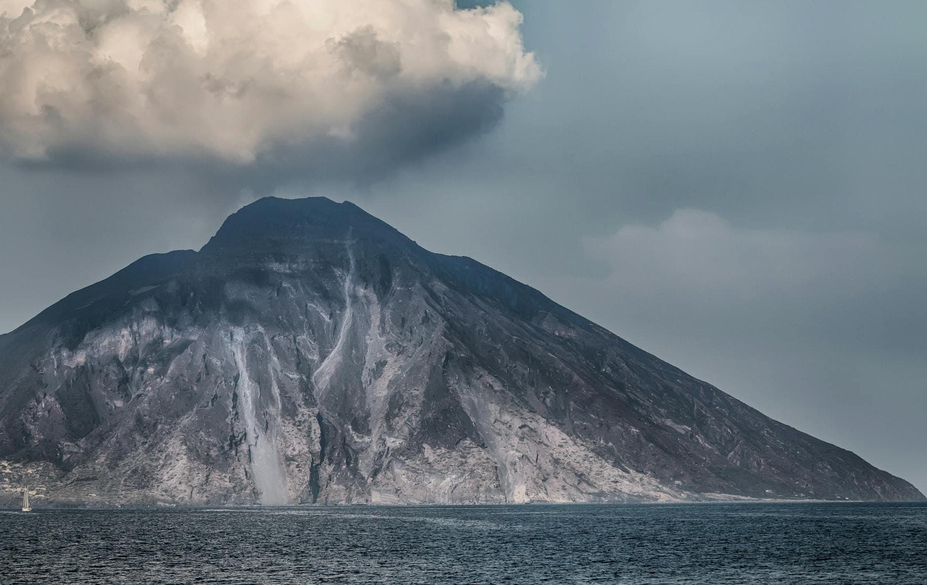 Pemandangan dramatis Gunung Berapi Stromboli yang menjulang dari laut, dengan asap mengepul dari puncaknya di bawah langit berawan.