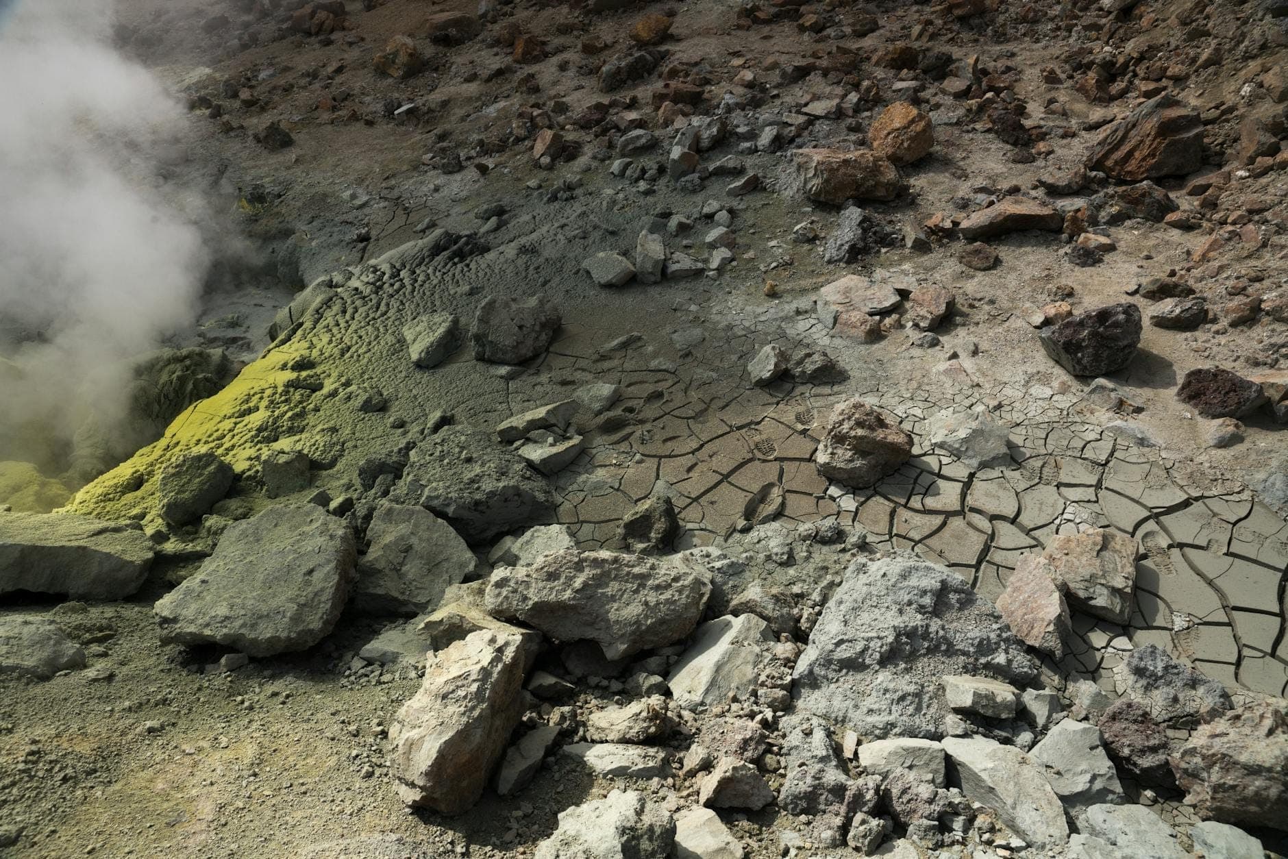 Dasar kawah vulkanik dengan endapan belerang, uap fumarol, dan lumpur kering yang retak di puncak gunung berapi.