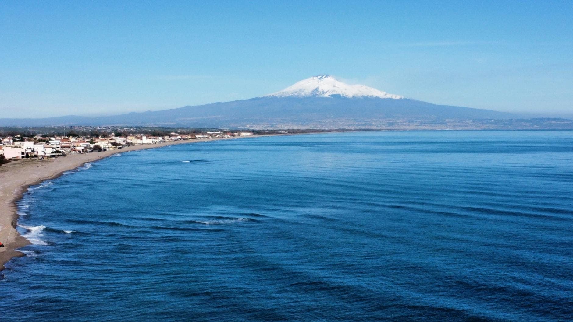 Gunung Etna bersalju menjulang di atas garis pantai Sisilia dengan pantai berpasir luas dan laut biru yang tenang di latar depan.