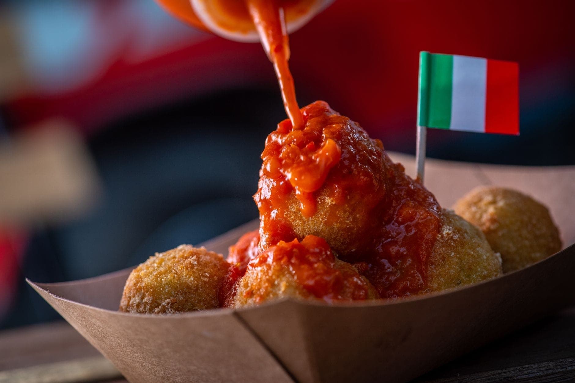 Foto close-up arancini dengan saus tomat yang dituangkan di atasnya, disajikan dalam nampan kertas dengan bendera Italia kecil.
