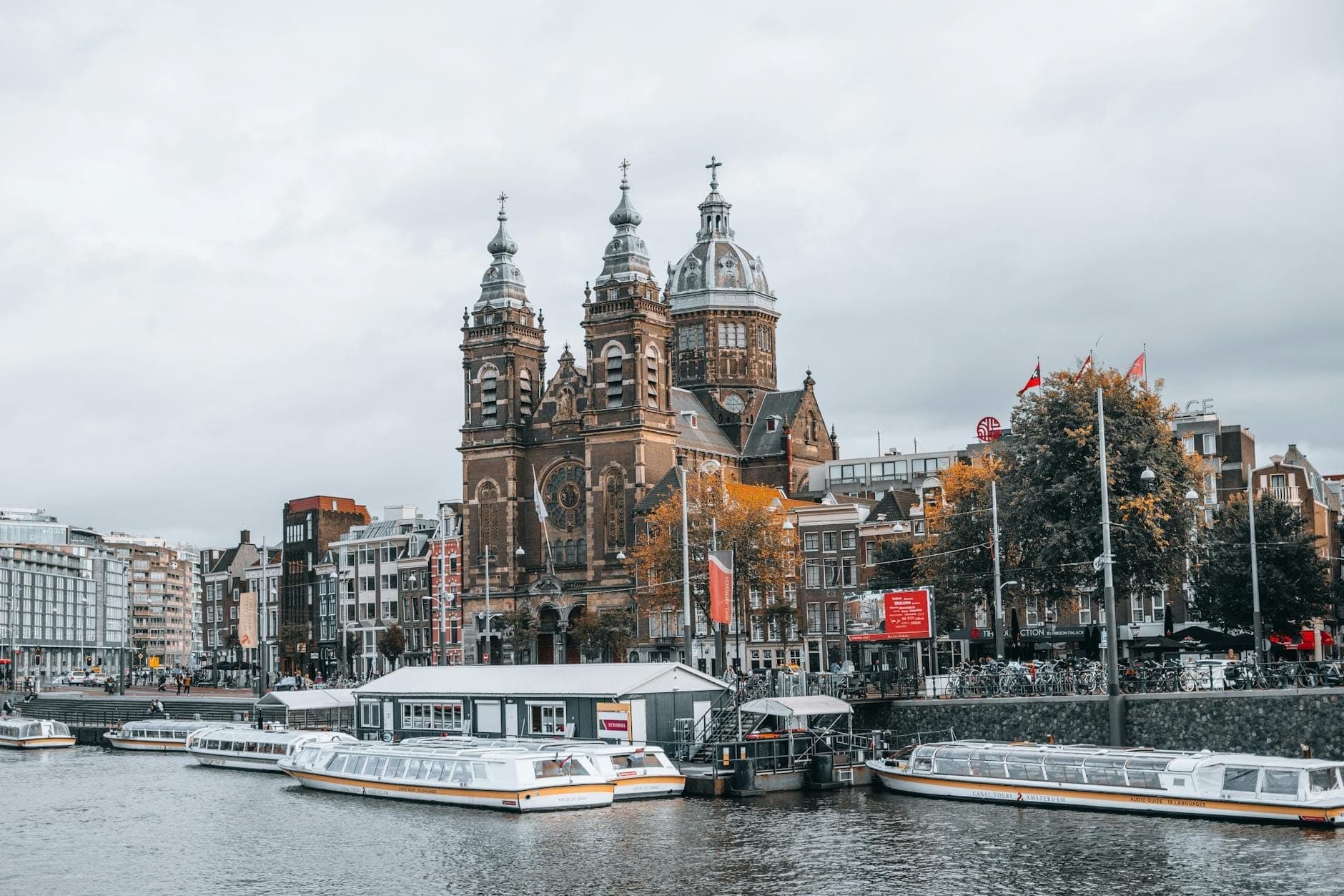 Vista amplia de la Basílica de San Nicolás en Ámsterdam con barcas en el canal en primer plano y los edificios de la ciudad bajo un cielo nublado.