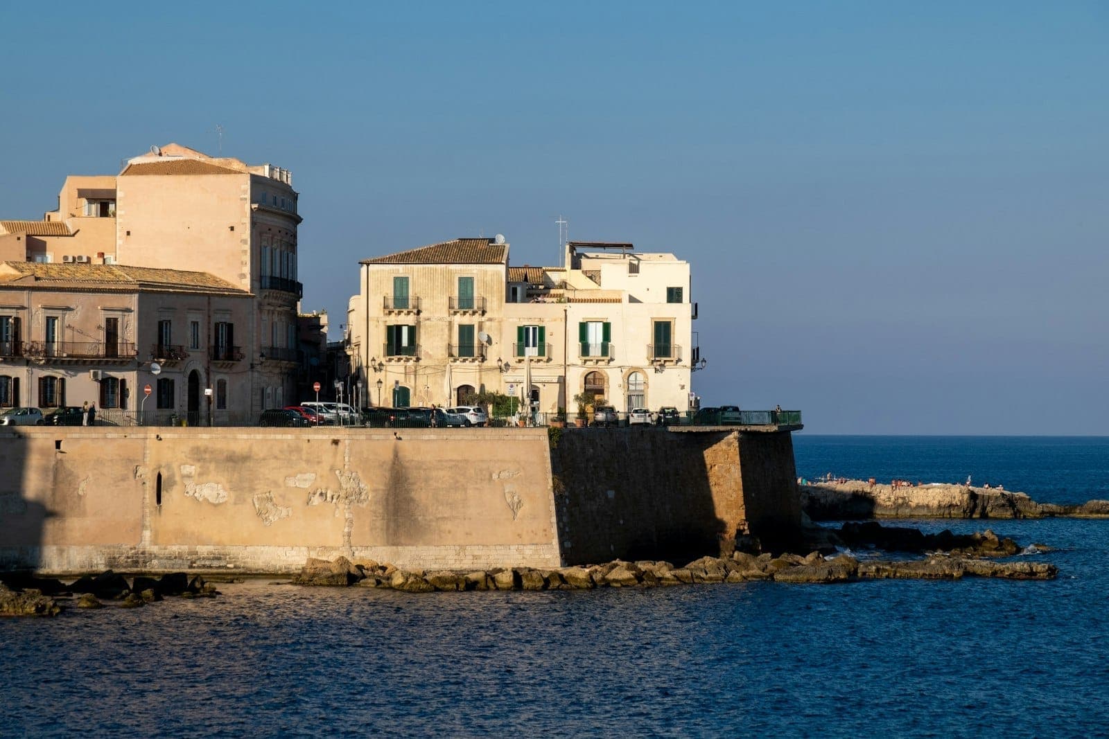 Pemandangan bangunan bersejarah di tepi air Ortigia, Siracusa, menjulang di atas tembok batu dengan Laut Mediterania biru di latar depan pada hari yang cerah.