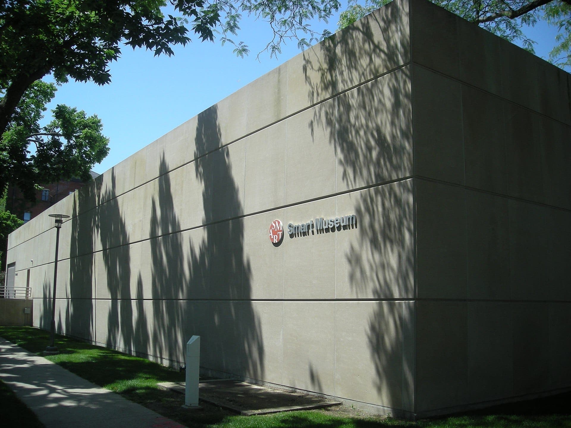 Exterior view of the Smart Museum of Art building at the University of Chicago, featuring minimalist concrete walls with tree shadows cast in sunlight.