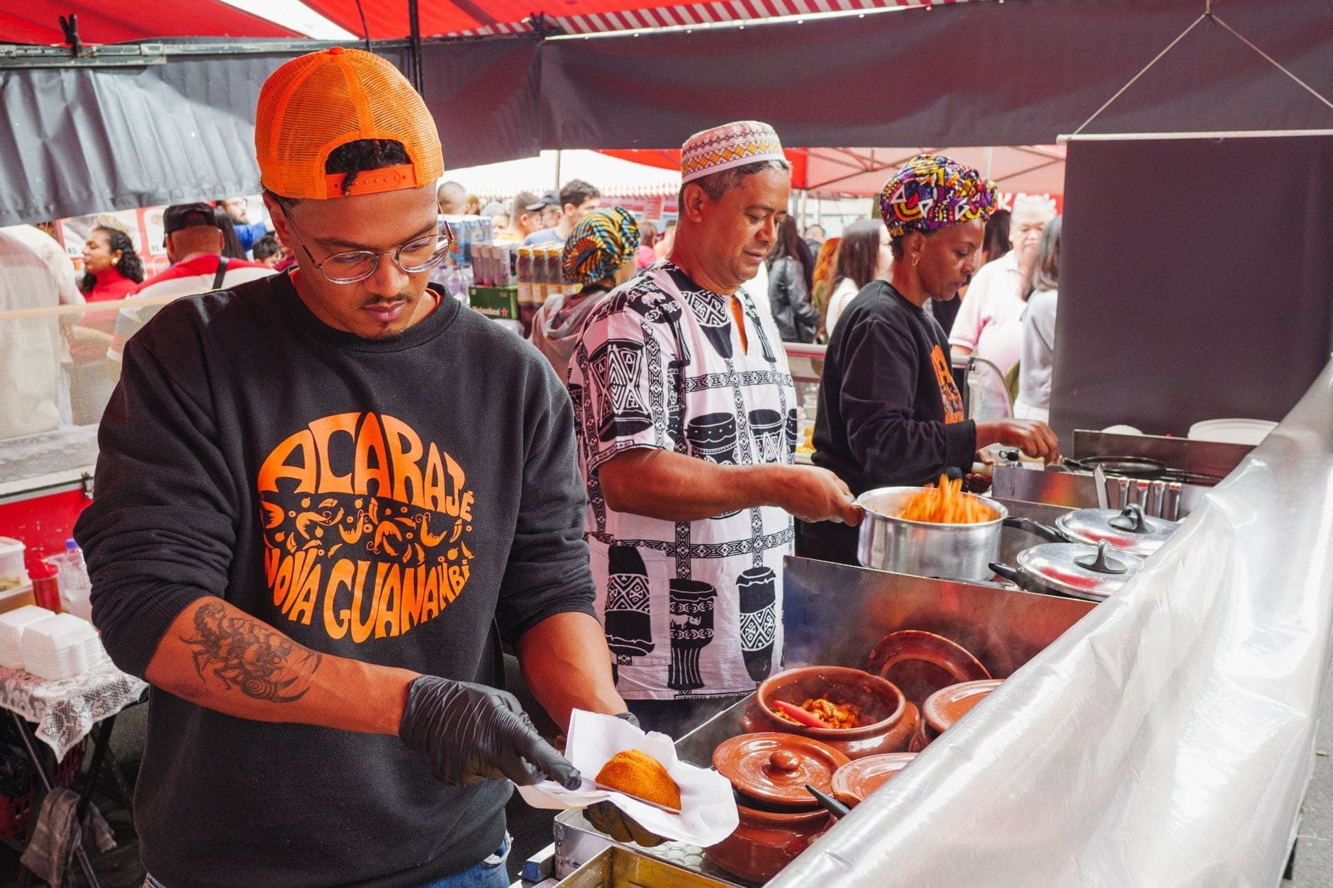 ผู้ขายเตรียมและเสิร์ฟอาหารที่แผงขายของสีสันสดใสในงาน Smorgasburg ตลาดกลางแจ้งของ Brooklyn ใต้เต็นท์สีแดงพร้อมฝูงชนอยู่เบื้องหลัง
