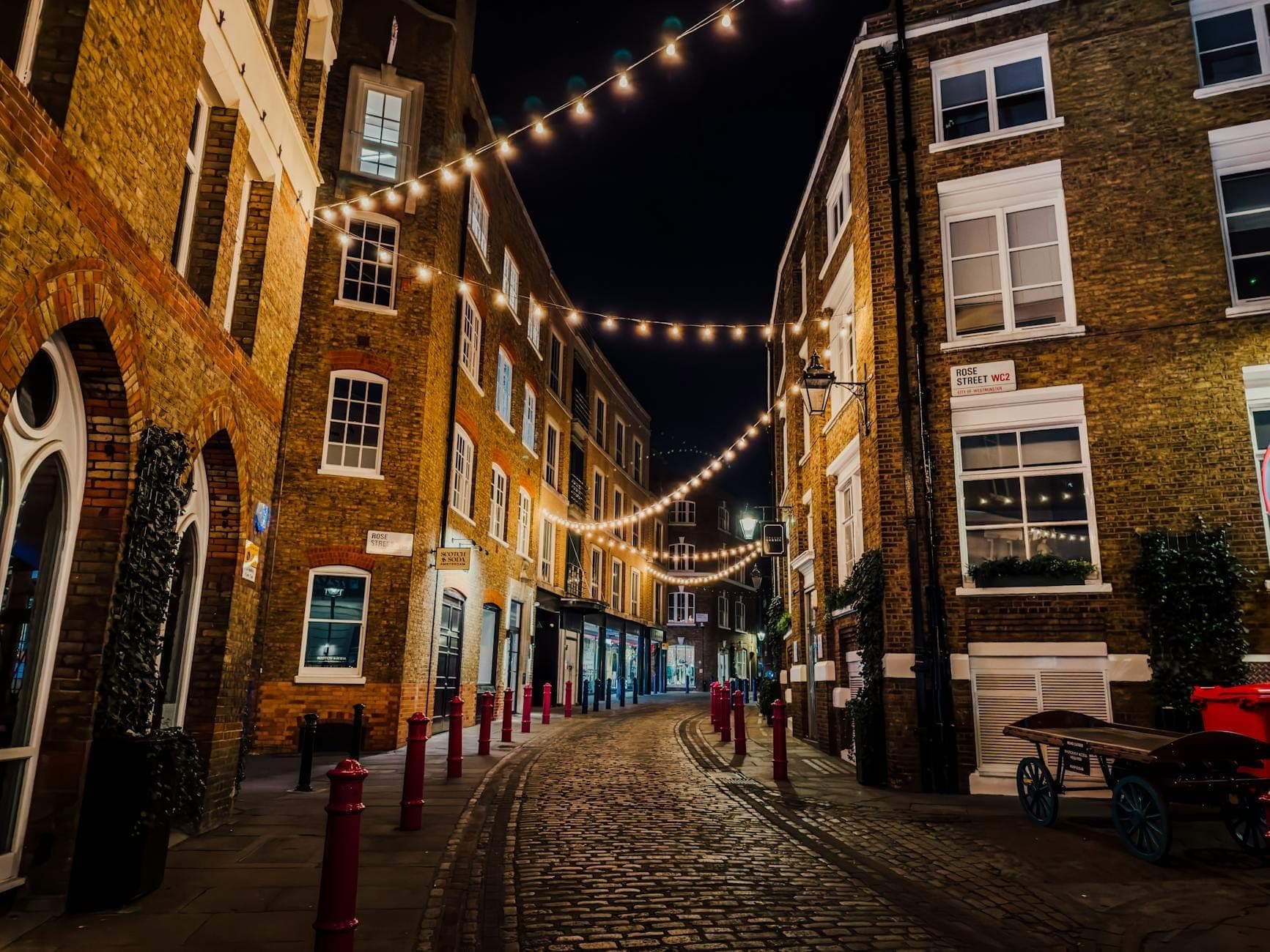 A charming cobbled street in Soho at night, lined with brick buildings and lit by warm string lights overhead, evoking the area’s vibrant after-dark energy.