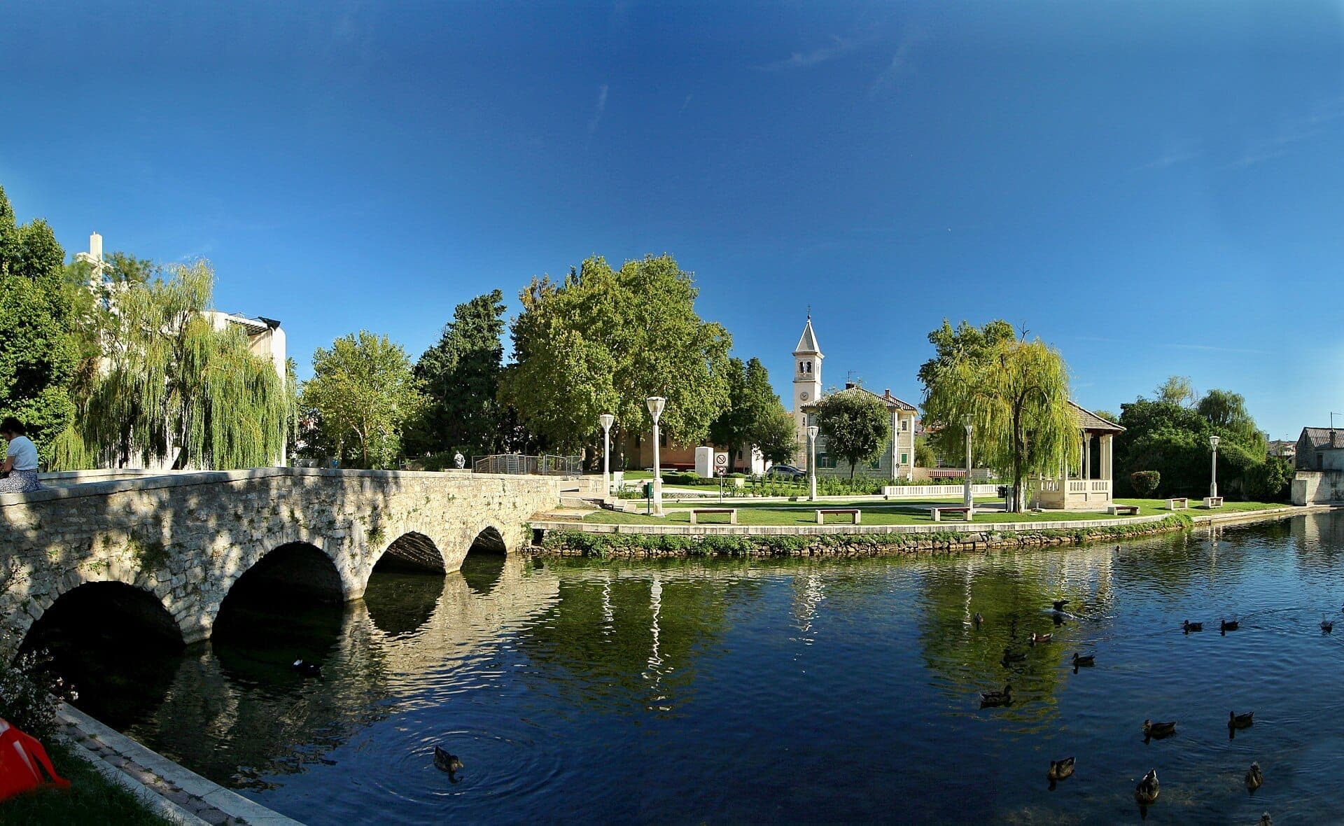 Ponte ad arco in pietra sul fiume Jadro a Solin, con rigogliosa vegetazione, anatre che nuotano e un campanile sotto un cielo azzurro brillante.