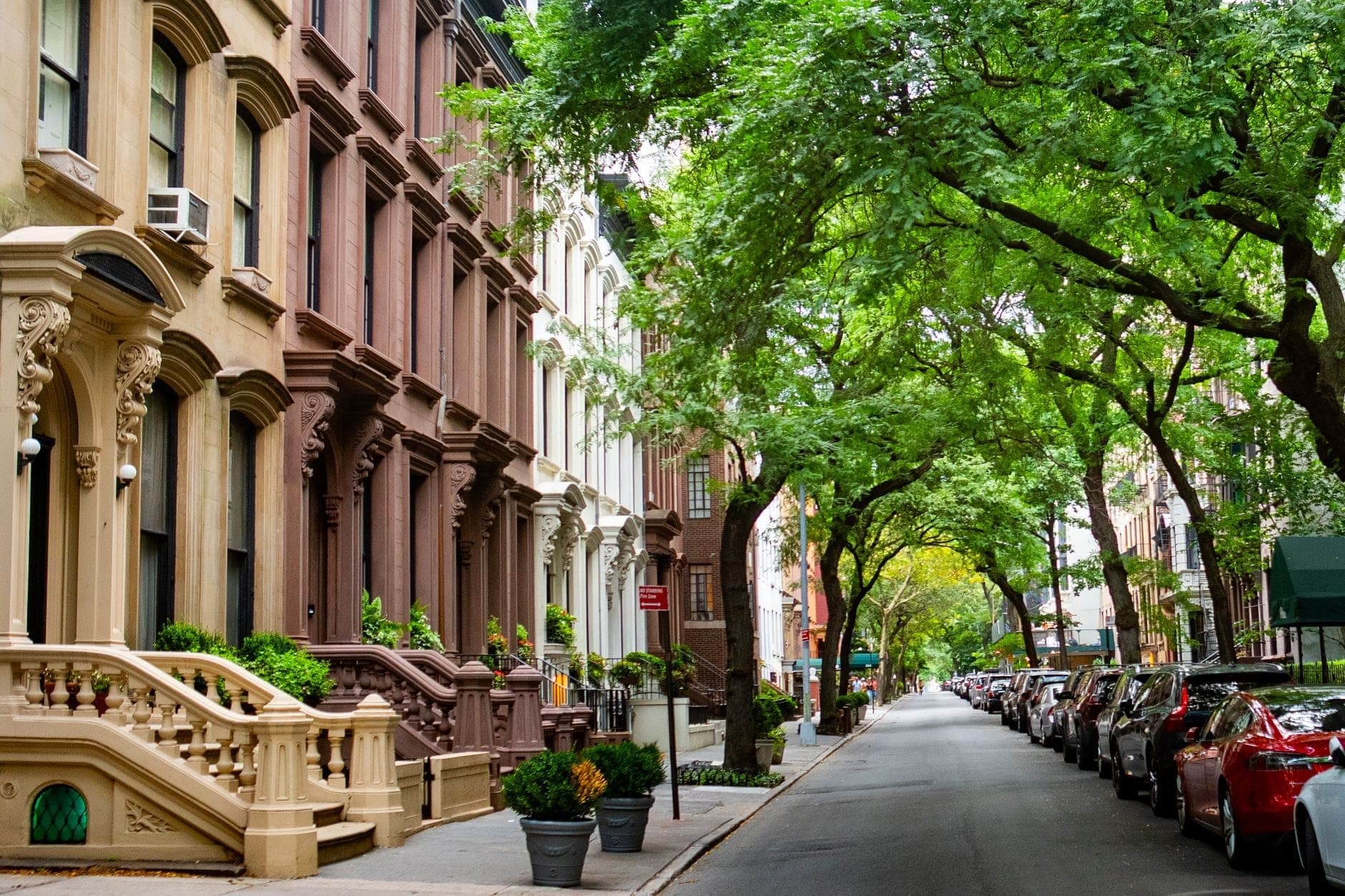 ถนนเรียงรายด้วยต้นไม้ใน New York City พร้อมบ้านแถวหินทรายสีน้ำตาลแบบคลาสสิกและรถจอดอยู่ สะท้อนบรรยากาศย่านที่พักอาศัย