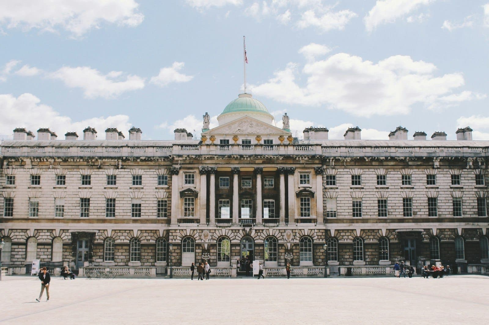 Die imposante neoklassizistische Fassade von Somerset House mit einer zentralen Kuppel, Steinsäulen und einem geräumigen Innenhof unter einem hellen, teils bewölkten Himmel.