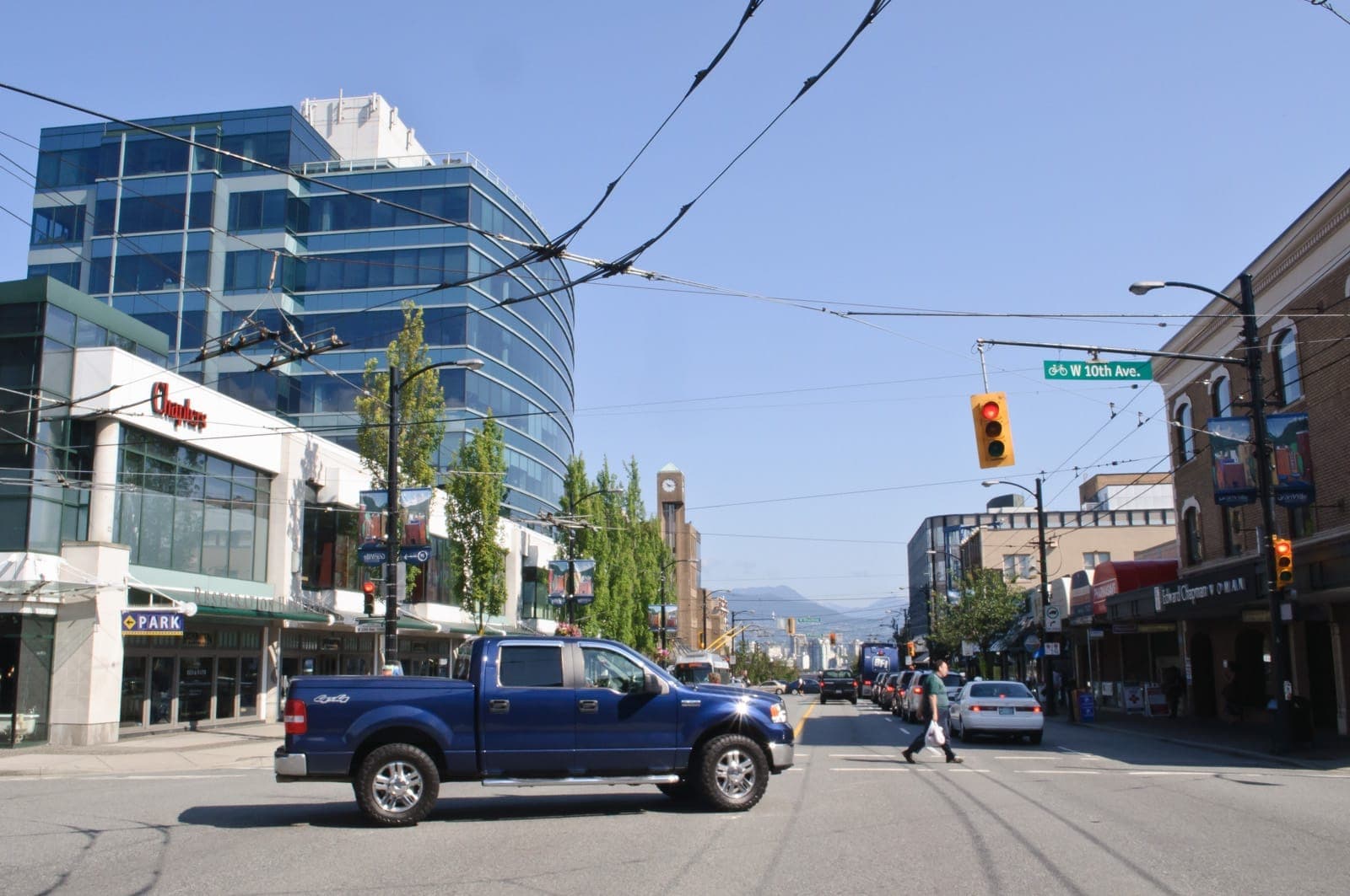สี่แยก South Granville และ West 10th Avenue ในแวนคูเวอร์ มีอาคารกระจกสมัยใหม่ ร้านค้า รถกระบะสีน้ำเงิน และท้องฟ้าสีฟ้าแจ่มใส